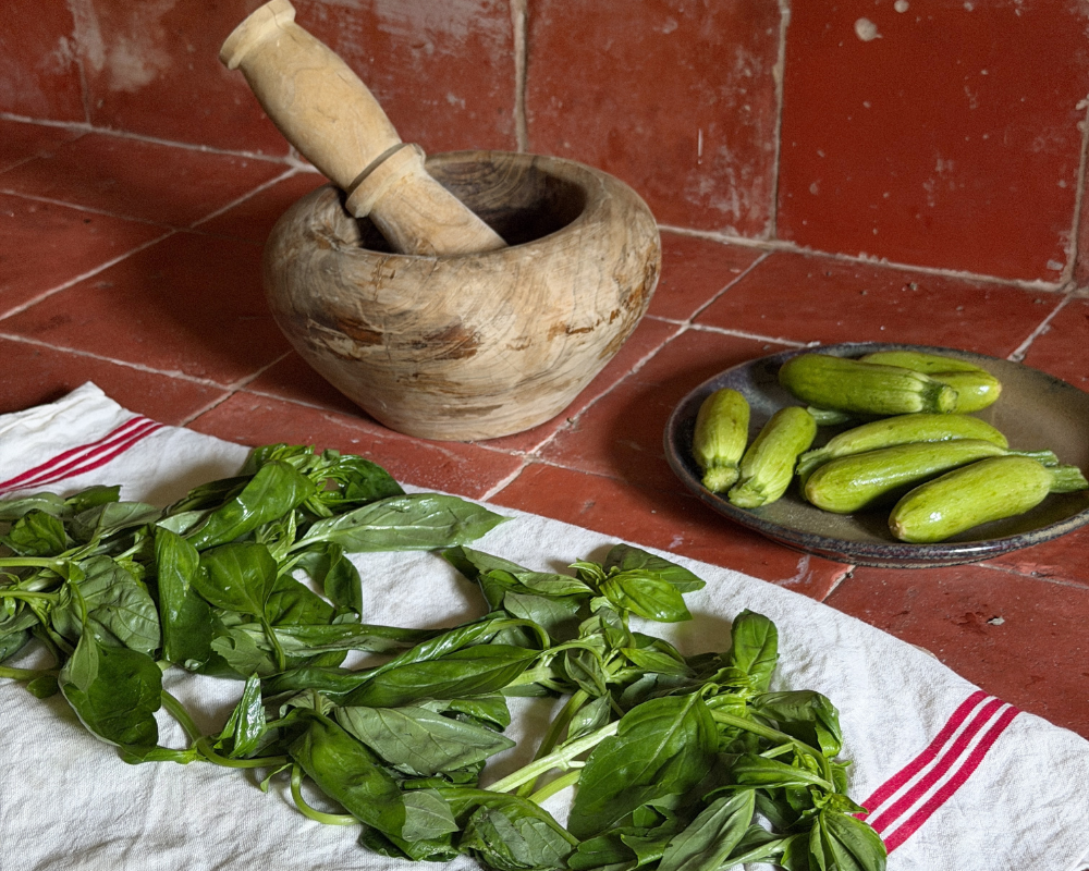  Fresh basil leaves on a linen cloth, with baby zucchini and a mortar and pestle in the background. 