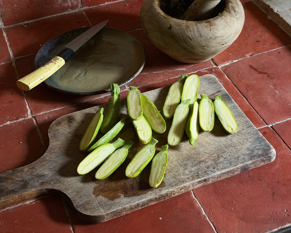  Halved baby zucchini arranged on a wooden board, with a knife and mortar and pestle nearby. 