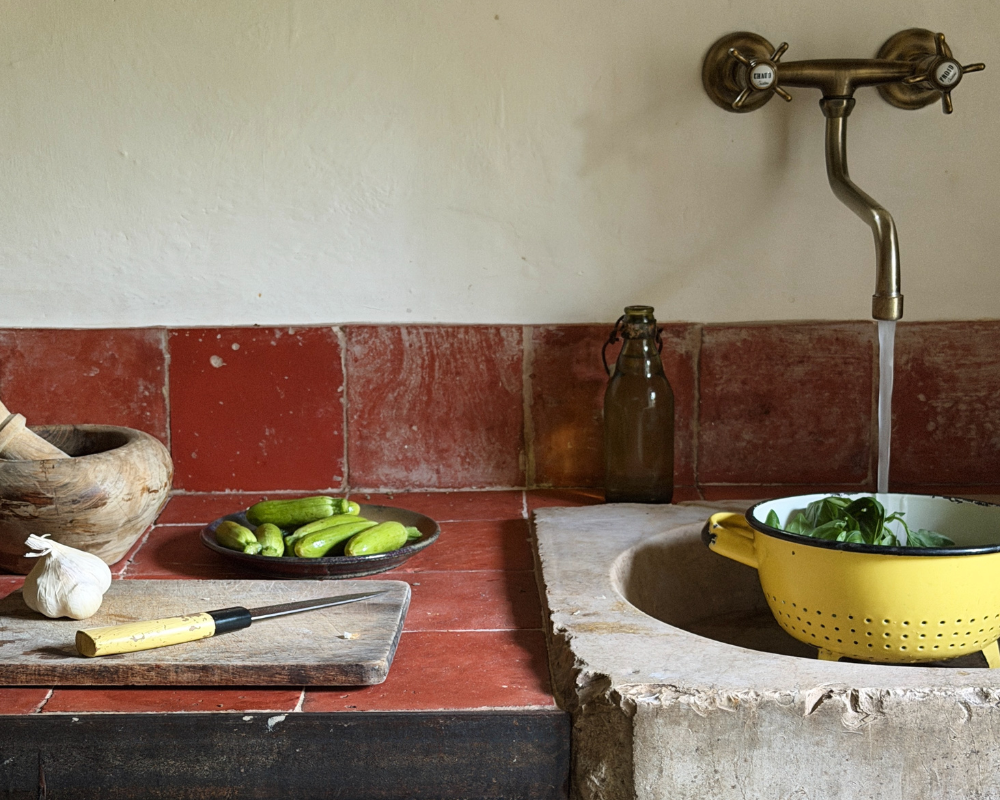  A rustic stone kitchen counter with baby zucchini, garlic and basil being rinsed in a yellow colander. 