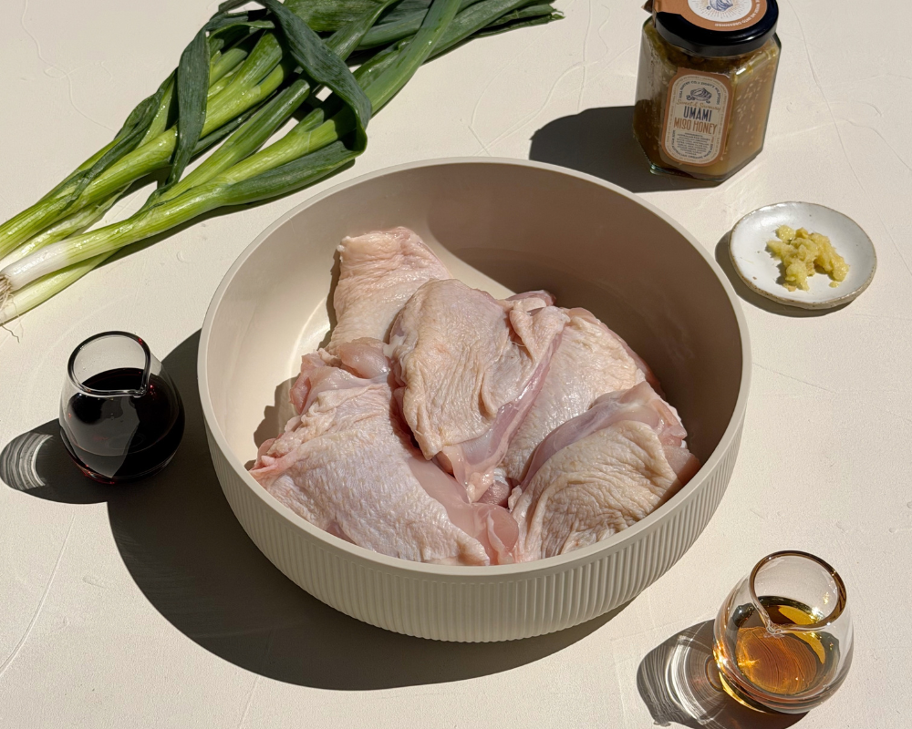  Raw chicken in a bowl with soy sauce, sesame oil, spring onions, ginger and miso honey around it for Sticky Miso Honey Chicken. 