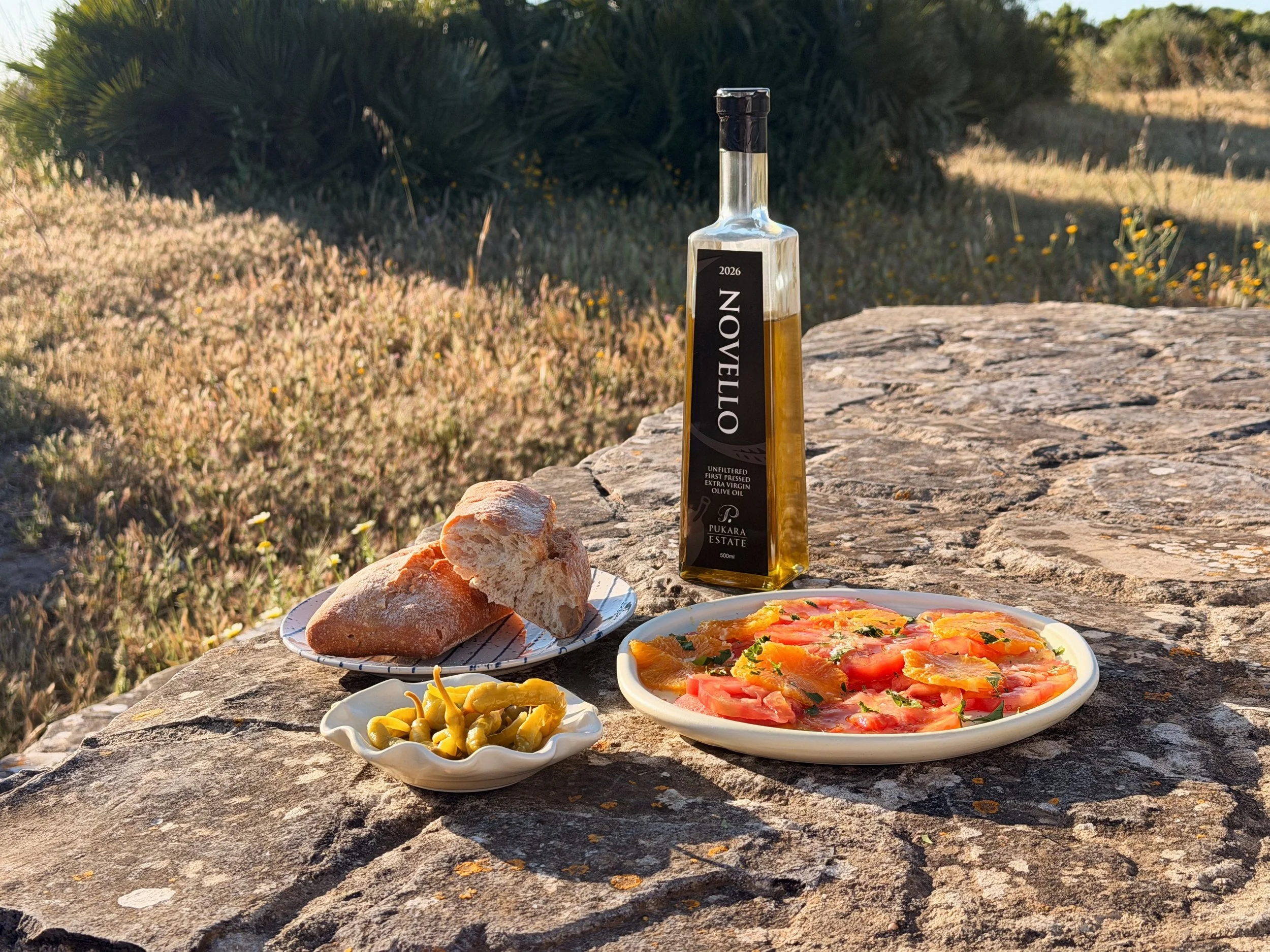 A bottle of Novello olive oil on a stone table next to a plate of bread, chillies and tomato and orange salad