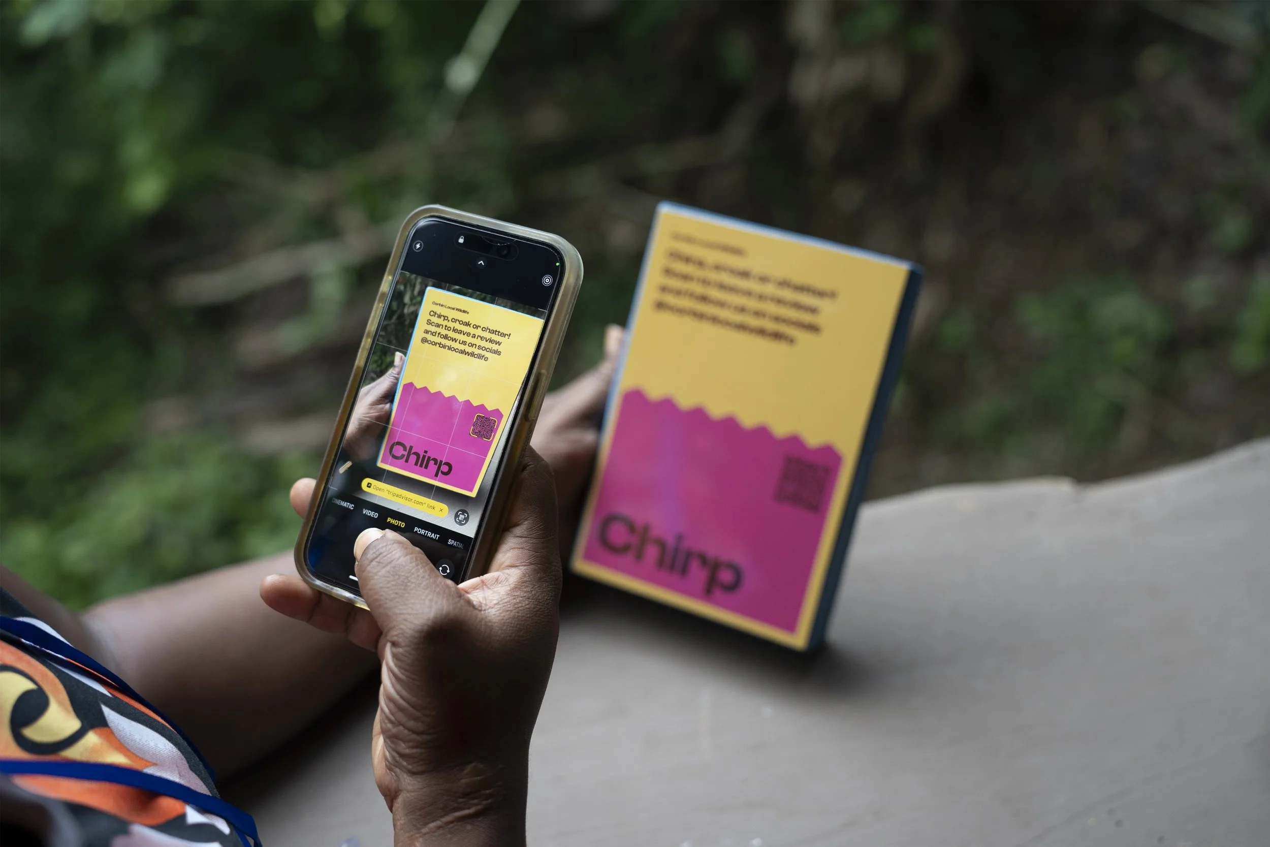 A person holding a smartphone taking a photo of a pink and yellow flyer with the word 'Chirp' on it, while sitting outdoors on a bench, with blurred greenery in the background.
