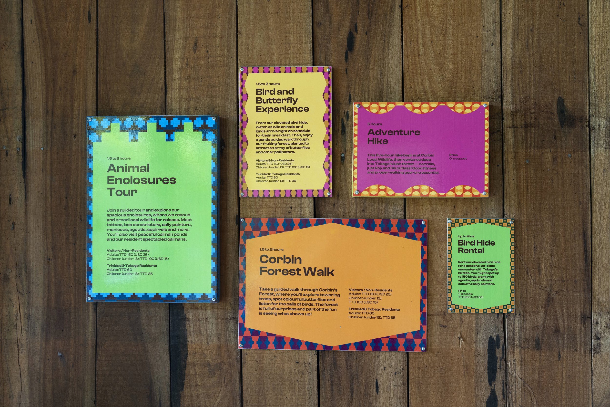 Colorful informational signs about a bird and butterfly experience, animal enclosures tour, corbin forest walk, adventure hike, and bird hide rental on a wooden wall.