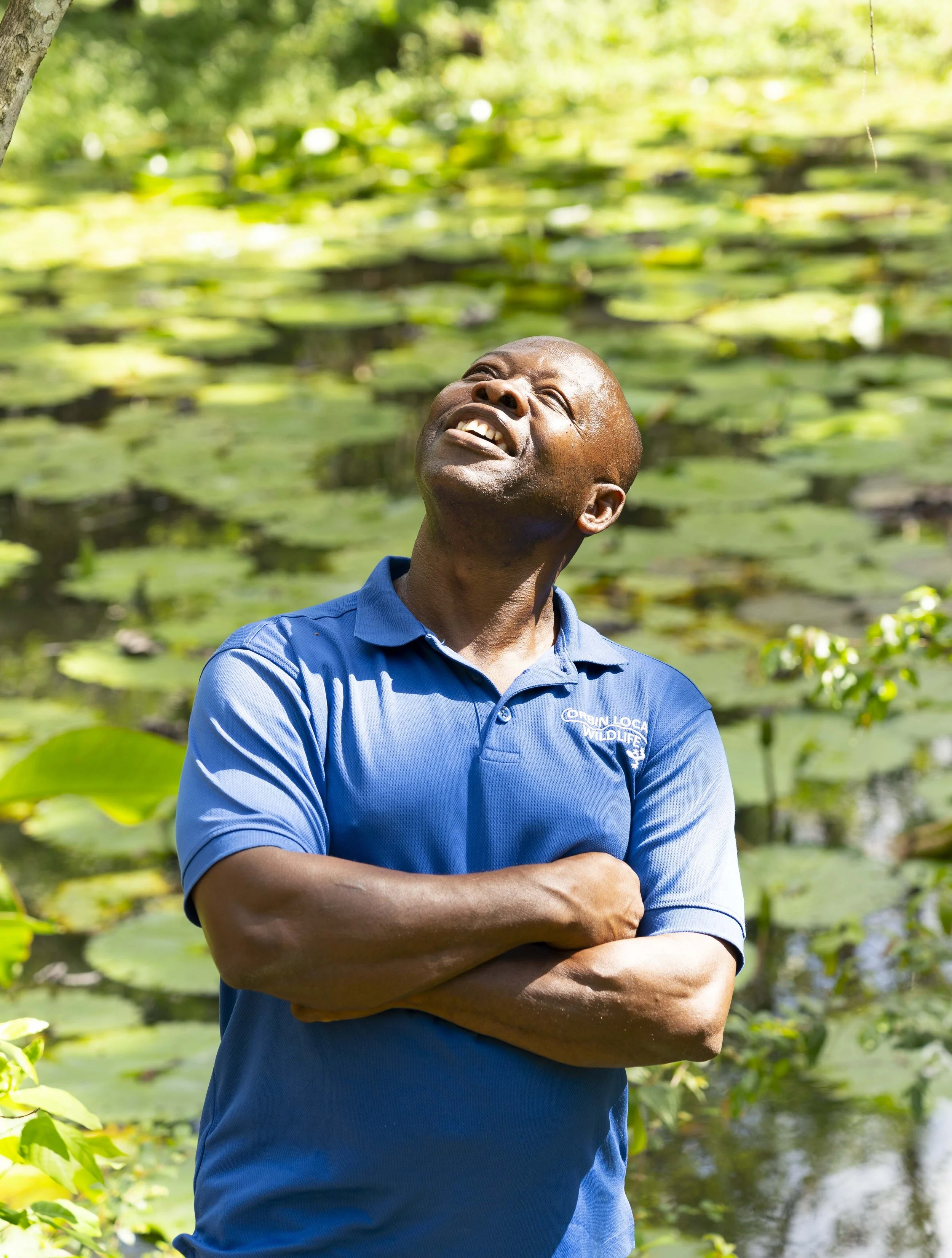 A man wearing a blue shirt with 'Orbin Local Wildlife' logo, arms crossed, looking upward with a thoughtful expression, standing outdoors near a pond with lily pads.