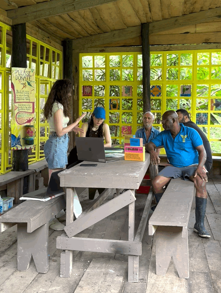 A group of people sitting and standing inside a wooden structure with large yellow-framed windows, discussing or listening to a young woman in casual clothes, with some using laptops and others attentively listening.
