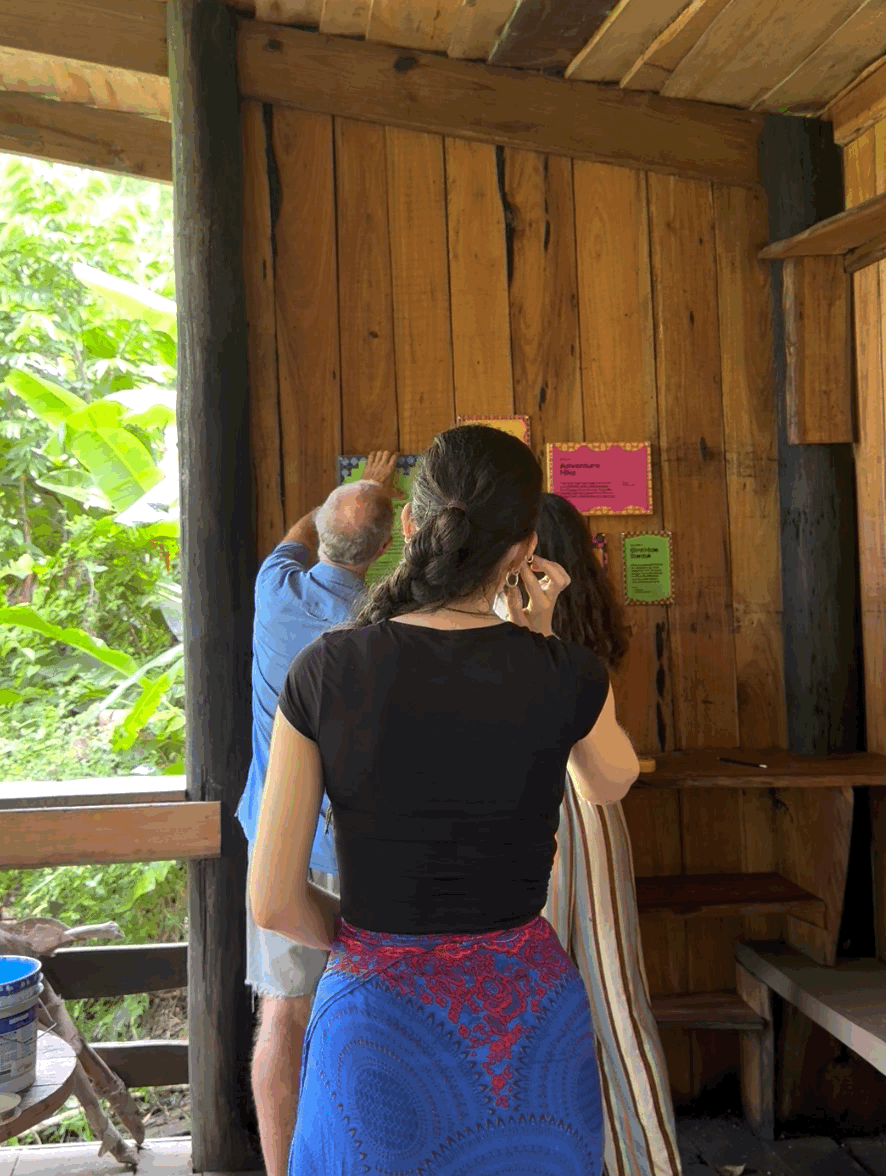 People viewing informational posters on a wooden wall inside a rustic wooden structure, with lush green foliage visible outside.