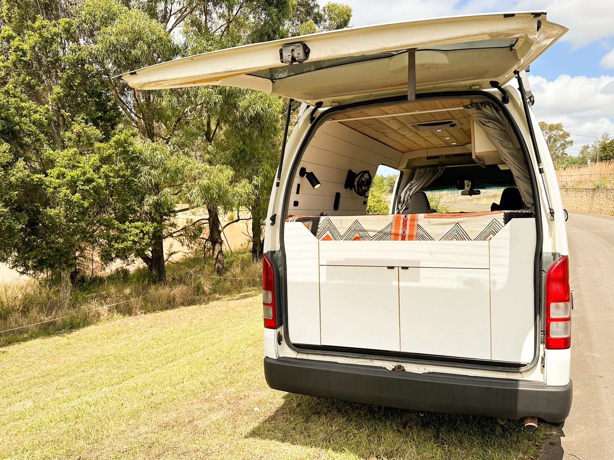 White van with its rear door open, parked on grassy area beside a road, with trees in the background.