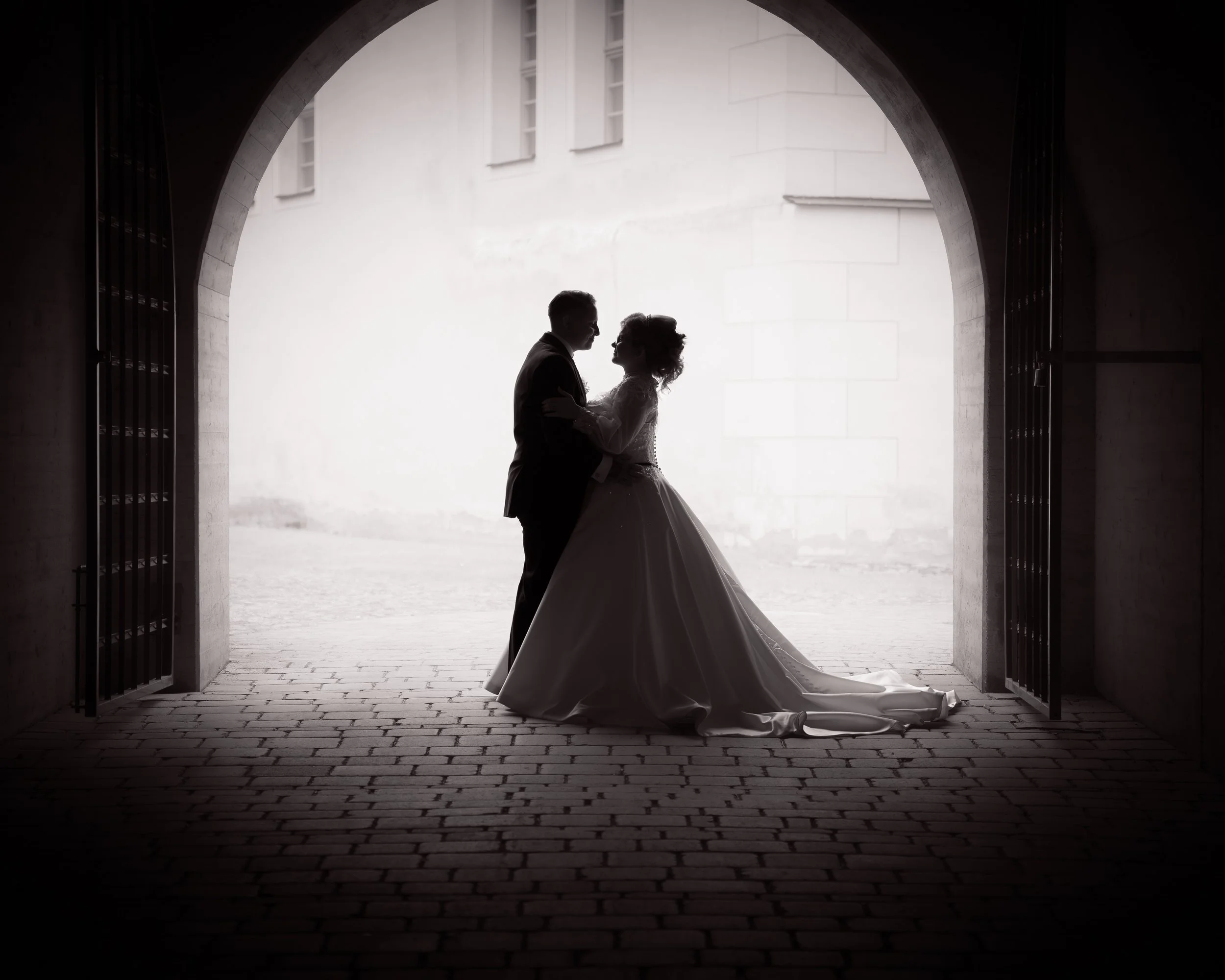 Silhouette eines hochzeitlichen Paares, das sich in einem Torbogen bei Sonnenuntergang oder Gegenlicht umarmt, mit einem historischen Gebäude im Hintergrund.