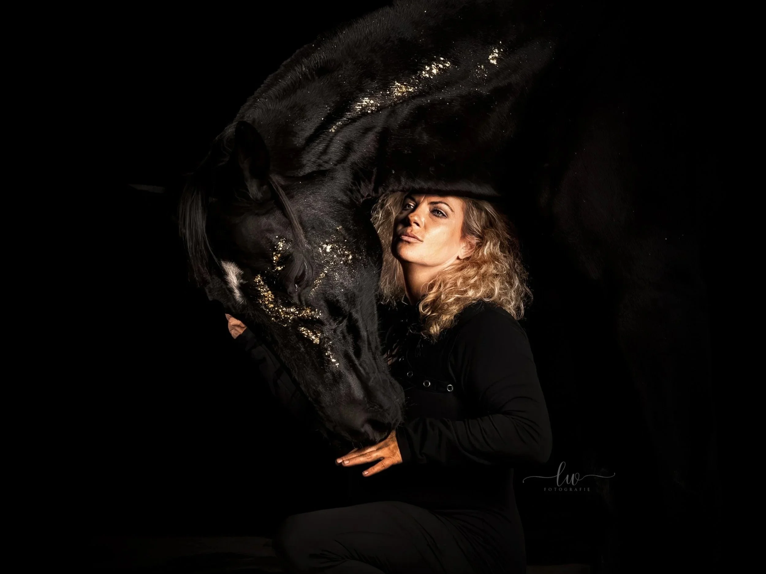 Eine Frau mit lockigem, hellbraunem Haar umarmt einen schwarzen Pferd mit goldfarbenen Flecken auf dem Kopf und Hals vor einem schwarzen Hintergrund.