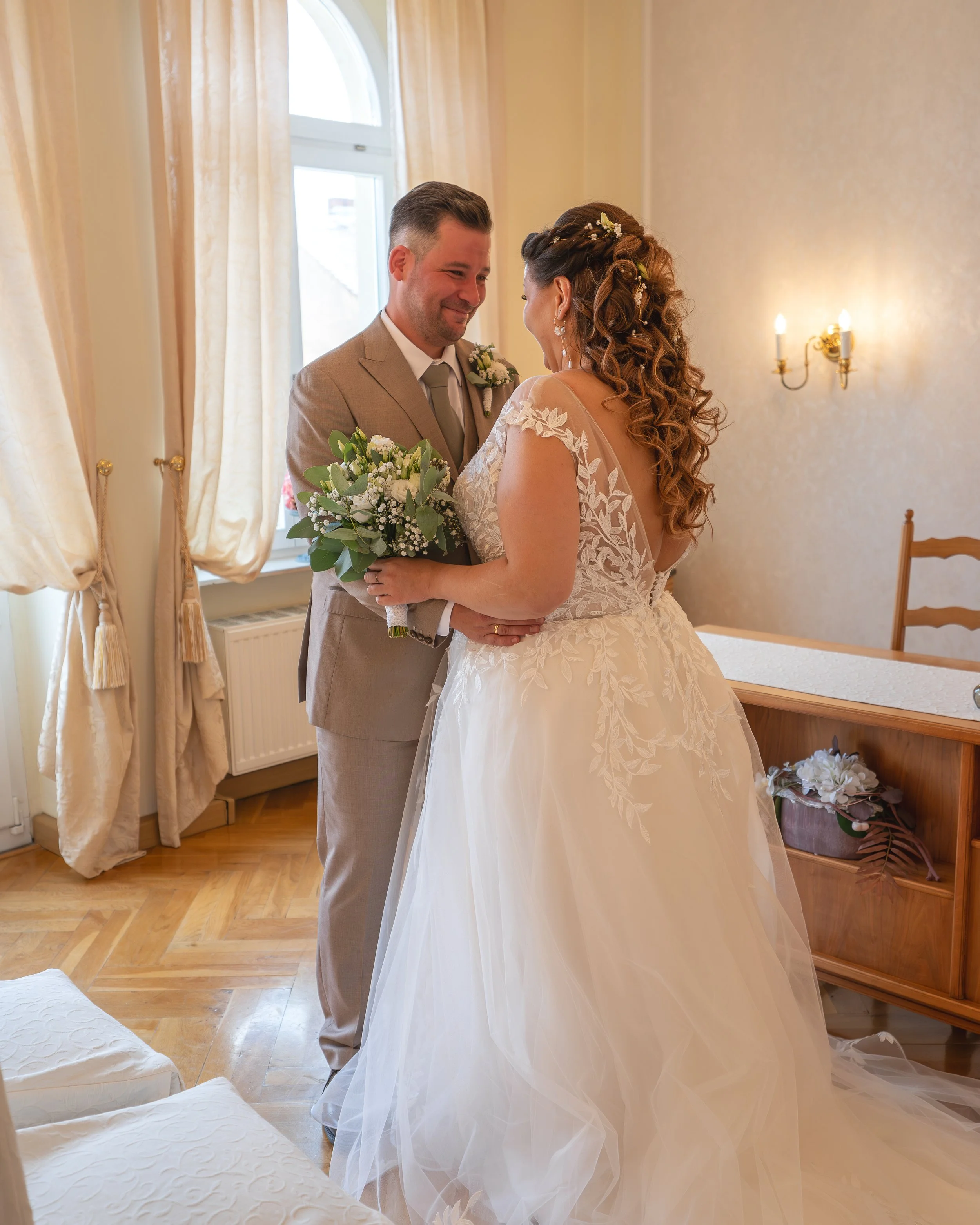 Ein Brautpaar blickt sich liebevoll an, steht in einem hellen, schön eingerichteten Raum mit Vorhängen an einem Fenster, die das Licht weich filtern, und einer Wandlampe. Die Braut trägt ein weißes, elegantes Brautkleid mit aufwendigen Stickereien und Haarschmuck, der Bräutigam einen hellgrauen Anzug mit Krawatte. Die Szene strahlt Liebe und Freude aus.