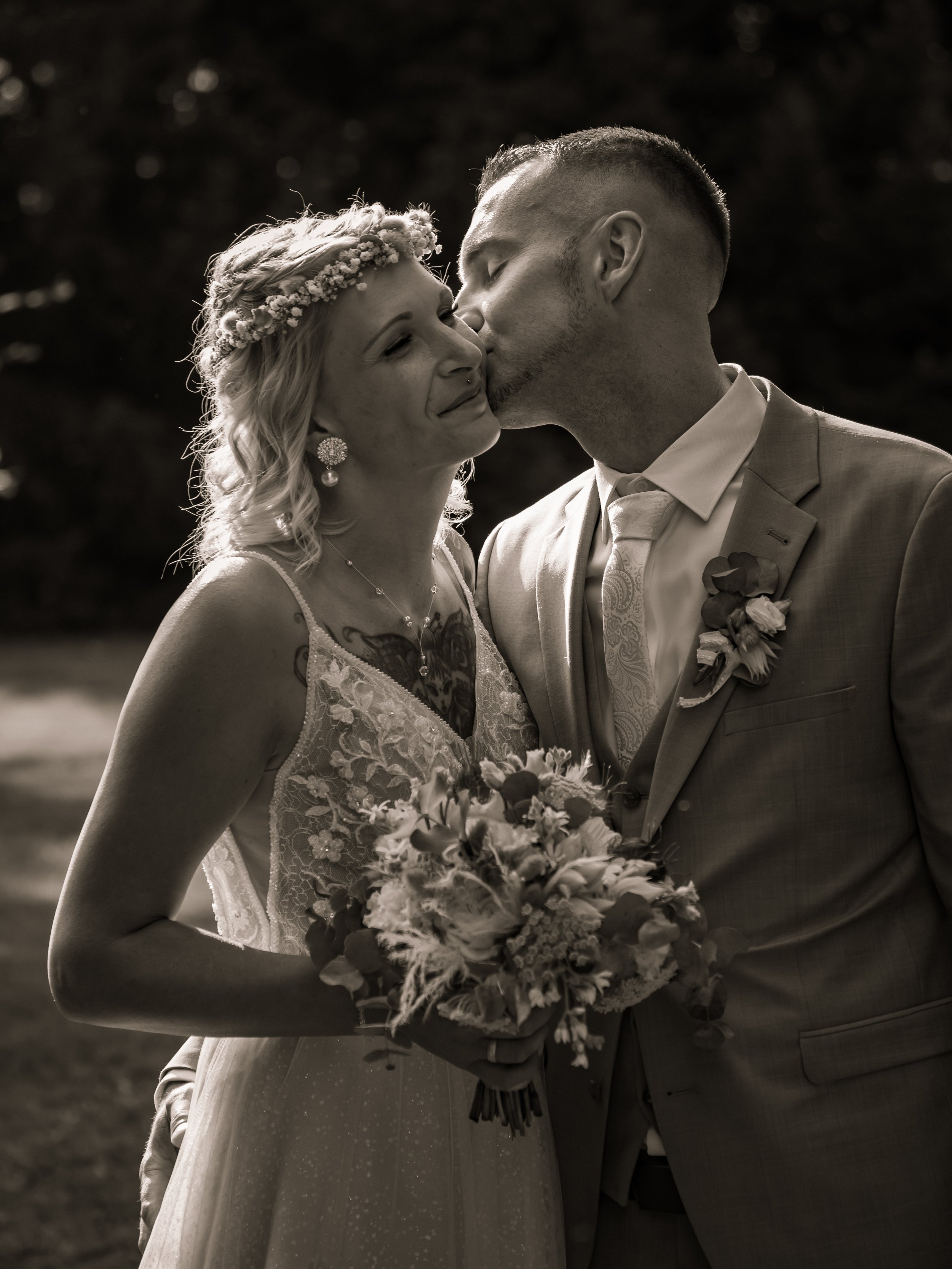 Ein braun-weißes Hochzeitsfoto eines Paares, das sich küsst, während die Frau einen Blumenkranz trägt und die Frau einen Brautstrauß hält, im Freien.