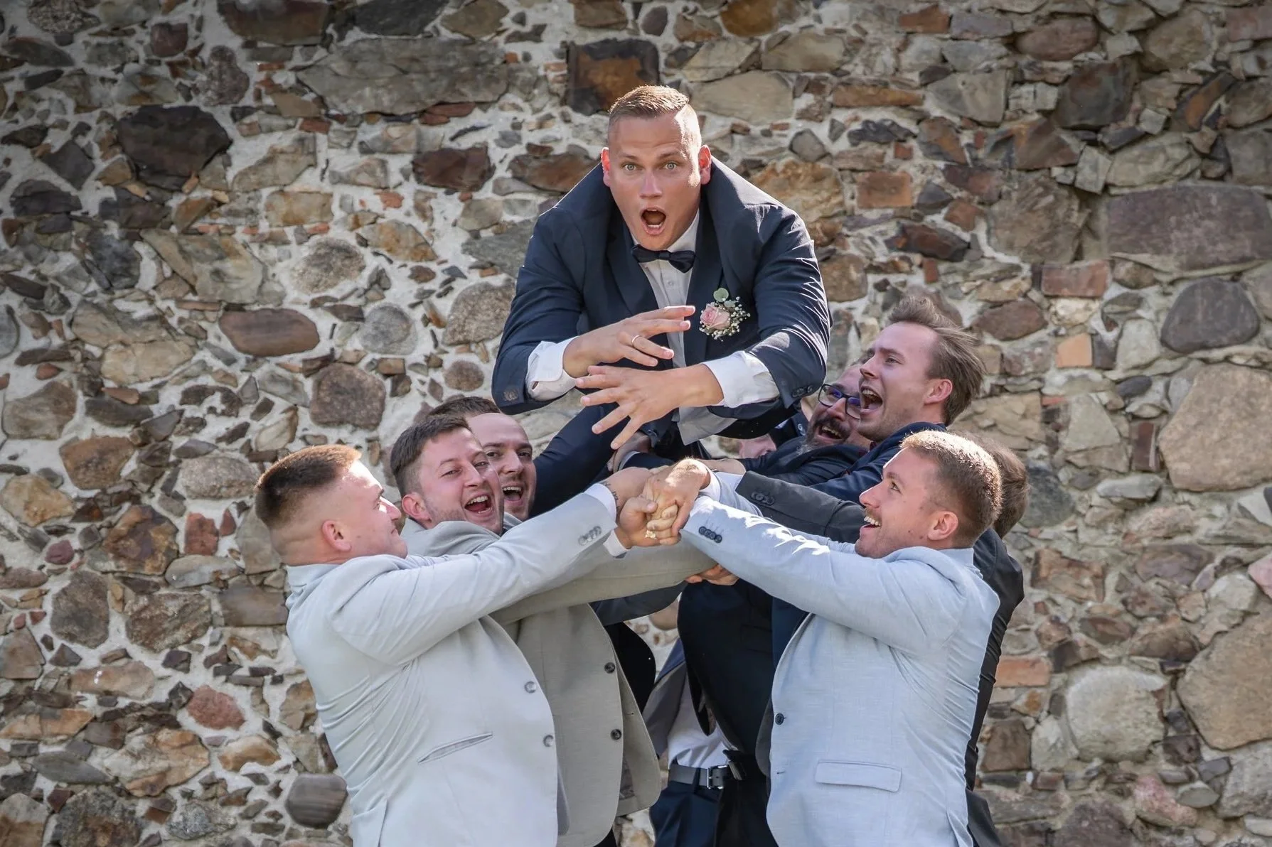 Eine Gruppe von Männern in Anzügen hebt einen Mann in Hochzeitskleidung hoch, während alle lachen und Freude zeigen.