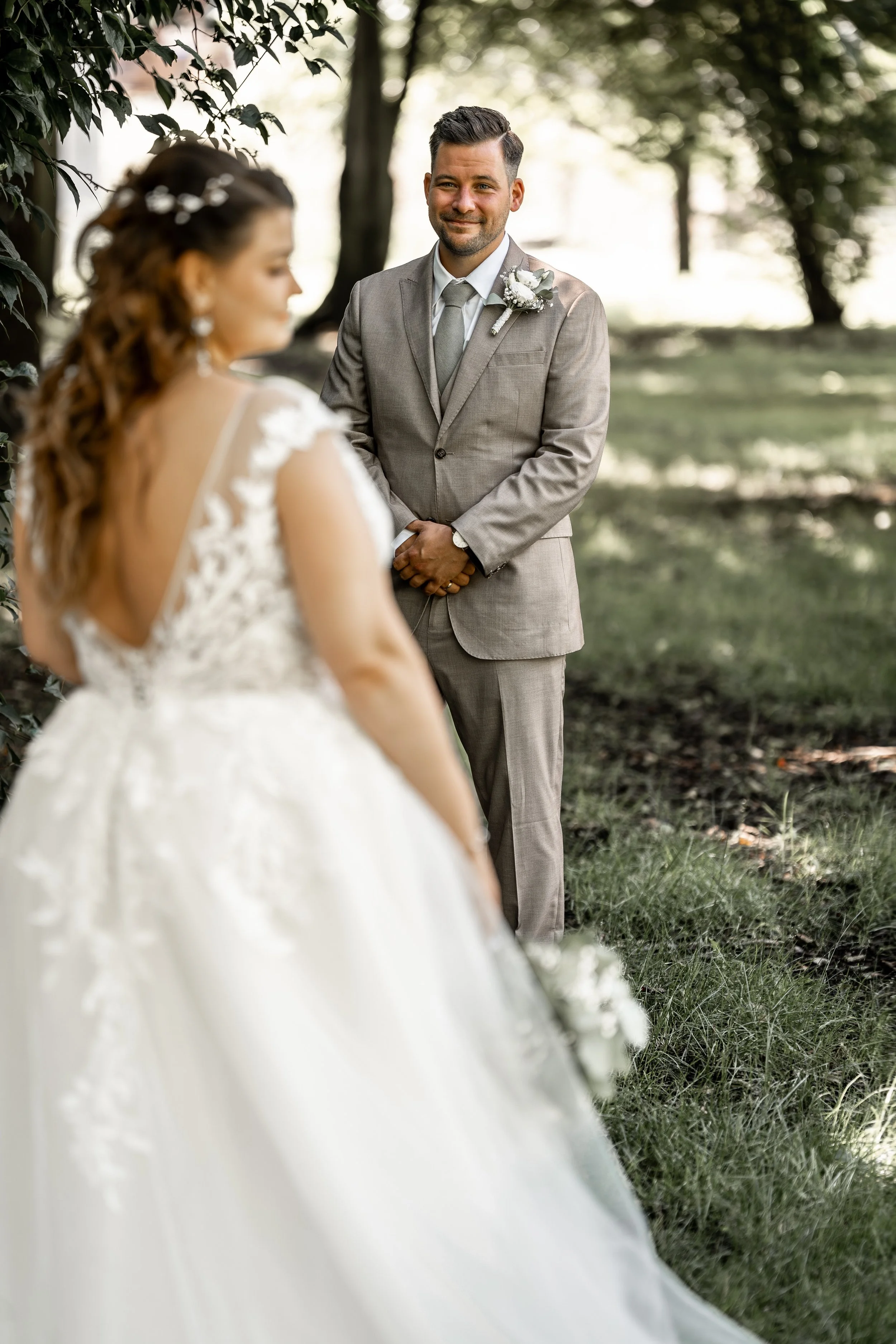 Ein Mann in einem grauen Anzug schaut auf eine Frau in einem weißen Brautkleid, die im Vordergrund unscharf ist, während sie im Freien steht, umgeben von Bäumen und Natur.