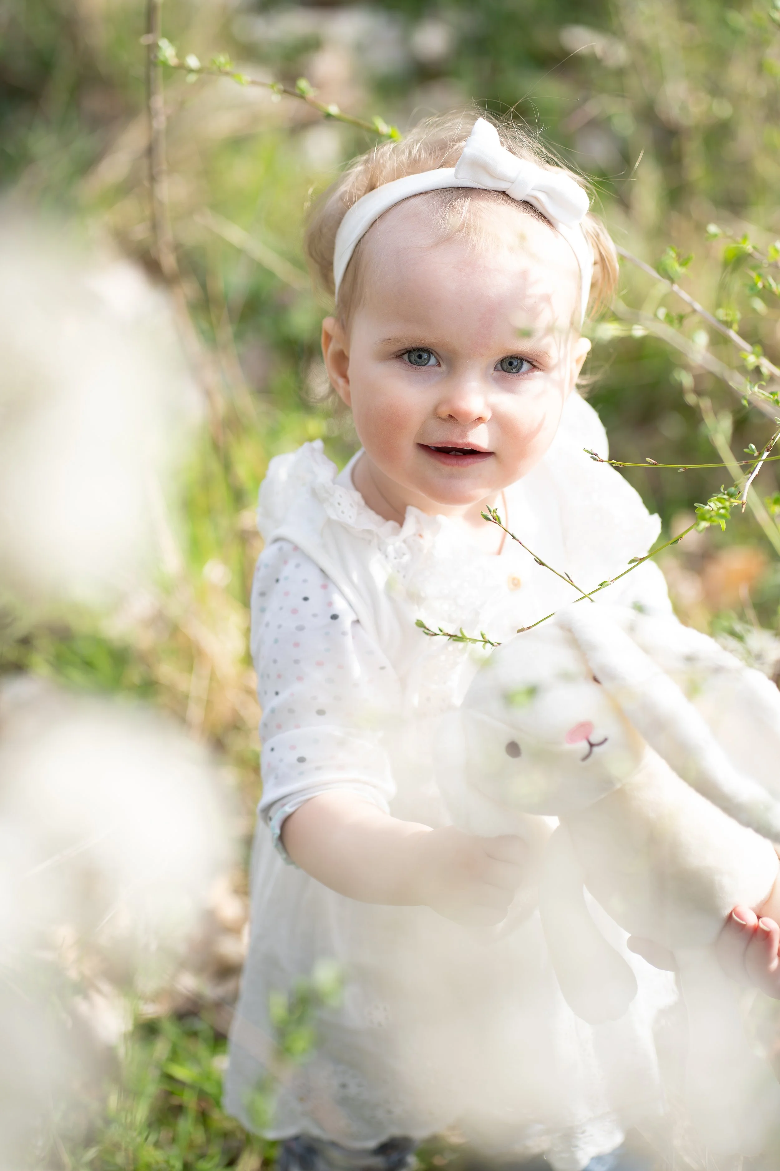 Ein kleines blondes Mädchen in weißer Kleidung mit rosa Punkten hält ein großes Kuscheltier im Außenbereich, umgeben von grünen Zweigen und blättern.