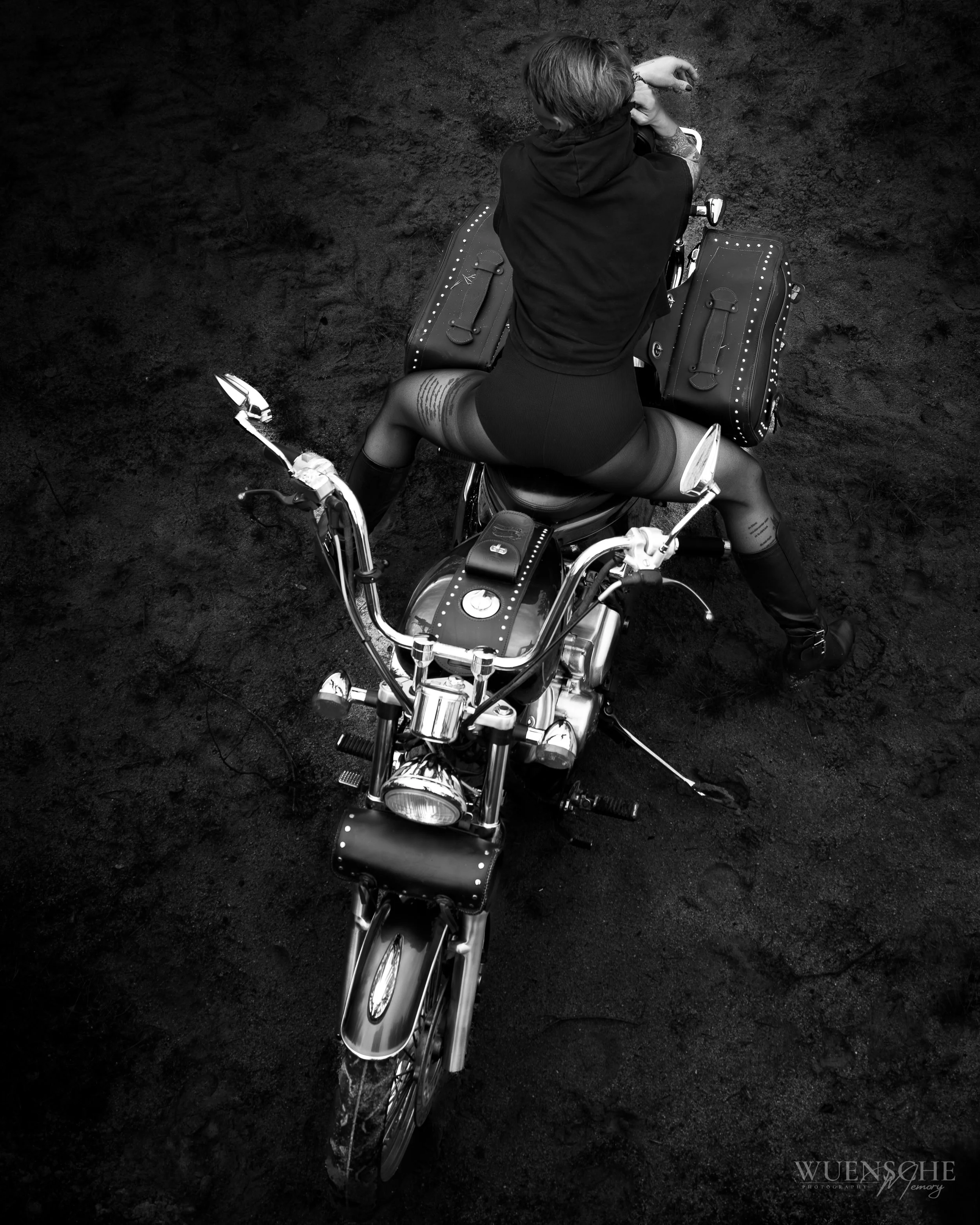 Ein Mensch sitzt rückwärts auf einem Motorrad auf dunklem Boden, trägt dunkle Kleidung und hat Tattoos auf den Beinen. Es sind zwei große schwarze Taschen am Motorrad befestigt. Das Bild ist in Schwarz-Weiß aufgenommen.