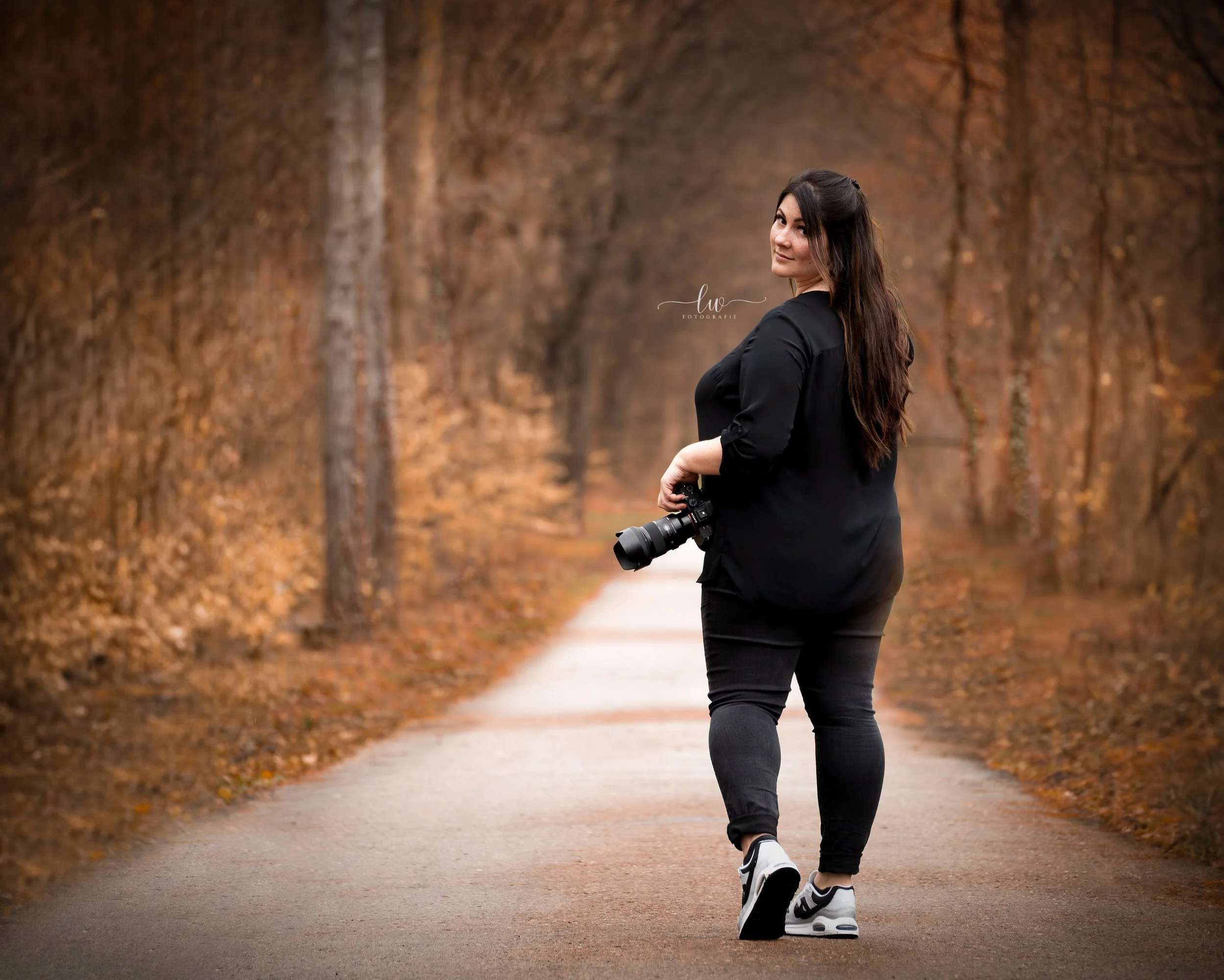 Eine junge Frau mit dunklem Haar trägt schwarze Kleidung und Turnschuhe, steht auf einem Weg in einem Herbstwald mit goldbraunen Blättern an den Bäumen, hält eine Kamera in der Hand und blickt lächelnd zurück.
