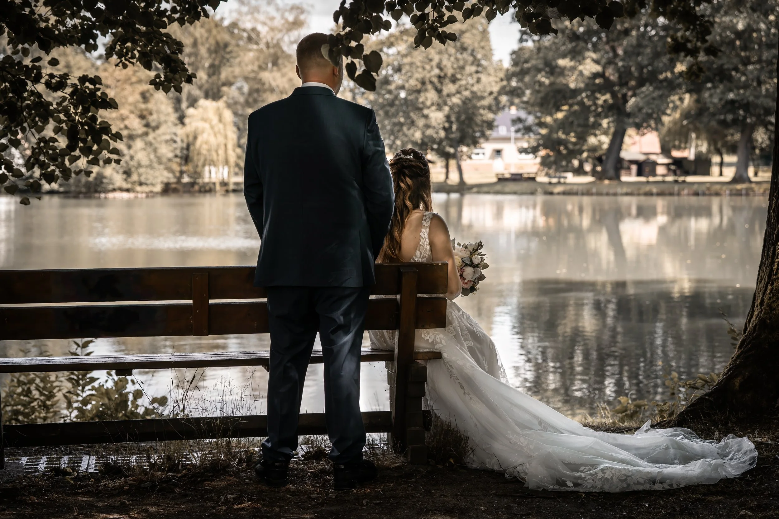 Ein Brautpaar bei einer Hochzeit am See, die Frau sitzt auf einer Bank in einem weißen Brautkleid mit Blumenstrauß, der Mann steht neben ihr in einem dunklen Anzug, der Blick ist zur Wasserfläche gerichtet, im Hintergrund sind Bäume und Häuser sichtb