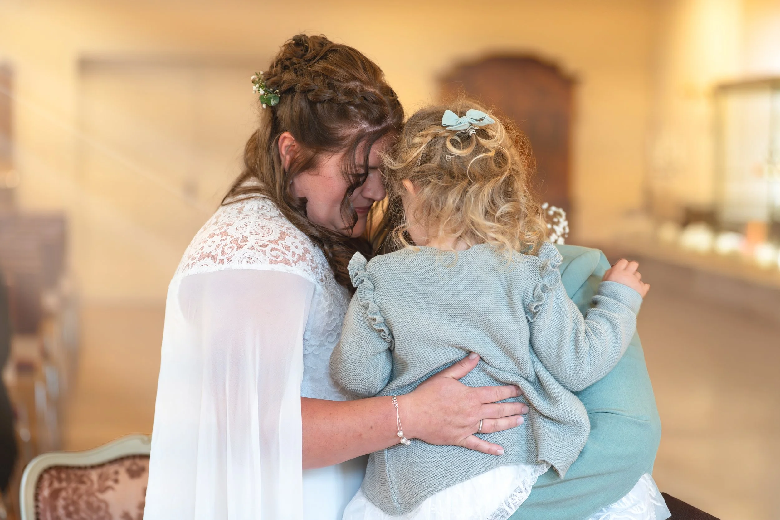 Zwei Frauen, die sich liebevoll umarmen, leicht ihre Stirnen aneinanderlegen, in einem hell erleuchteten Raum, möglicherweise bei einer Hochzeit oder Feier.