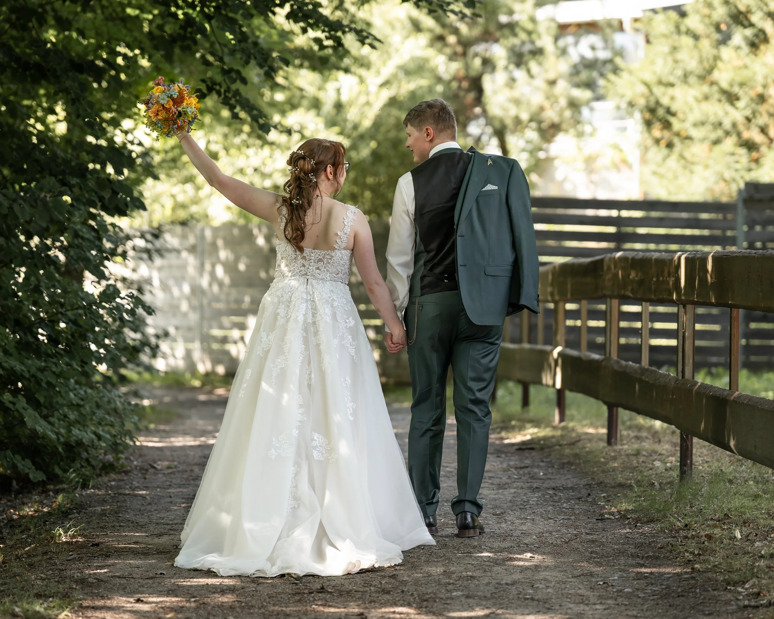 Ein Brautpaar geht Hand in Hand durch einen grünen Park, die Braut trägt ein weißes Hochzeitskleid und hält einen bunten Blumenstrauß, während der Bräutigam einen dunklen Anzug mit Weste trägt. Im Hintergrund sind Bäume und eine Holzzäune zu sehen.