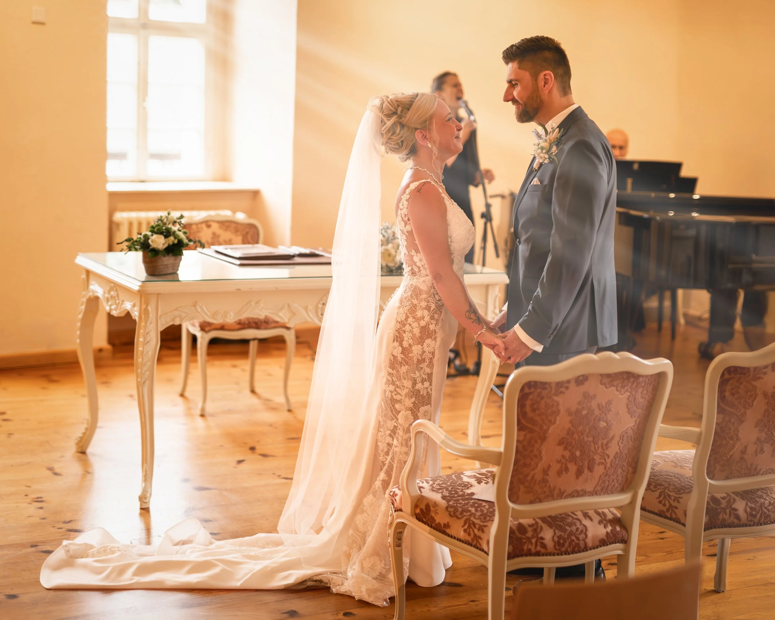 Ein Brautpaar hält sich bei ihrer Trauung im Standesamt liebevoll an den Händen, die Braut trägt ein cremefarbenes Spitzenkleid mit Schleppe, der Bräutigam einen Anzug mit Fliege. Im Hintergrund stehen Musiker, die Zeremonie findet in einem hell erleuchteten Raum statt, mit antiken Möbeln und einem Blumenstrauß auf einem Tisch.
