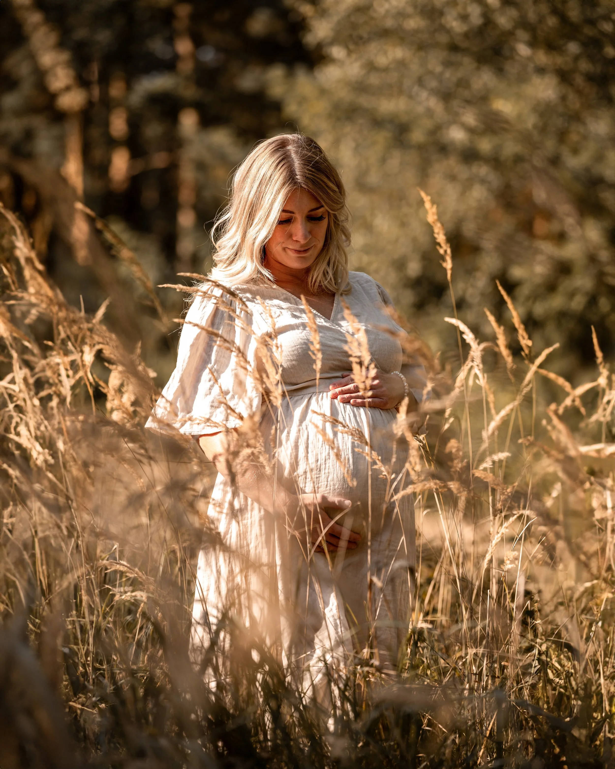 Eine Frau in einem hellen Kleid steht in einem goldfarbenen Getreidefeld und hält die Hände auf den Bauch, während sie nach unten schaut.