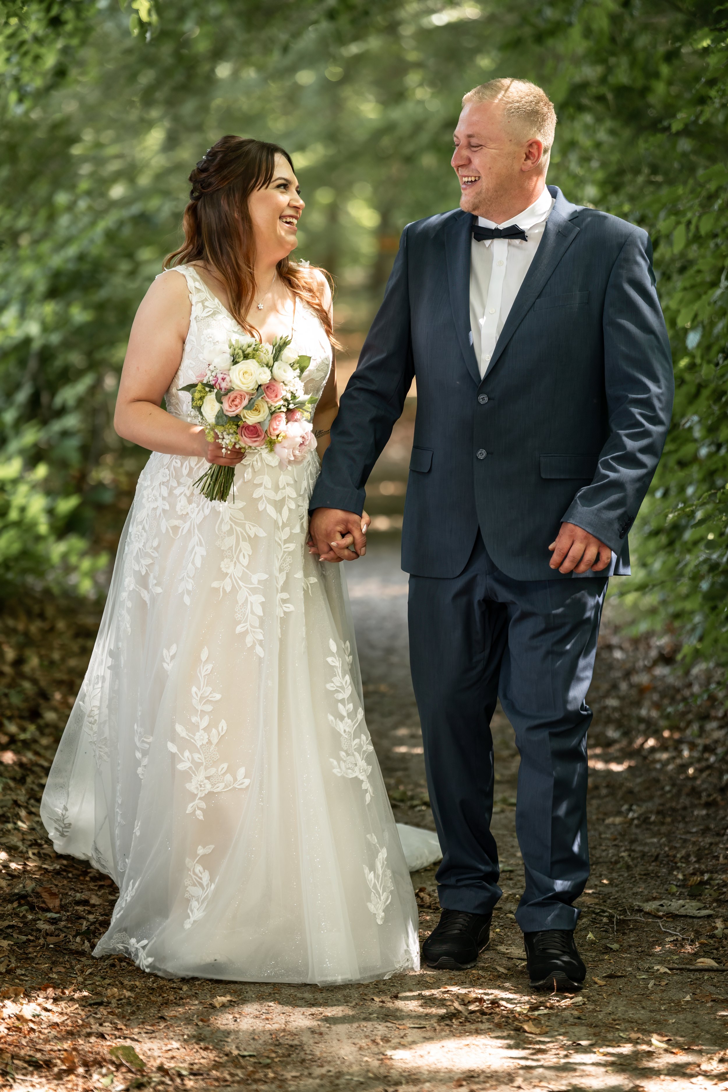 Ein lächelndes Brautpaar hält Händchen in einem Wald, sie trägt ein elegantes weißes Brautkleid mit Blumenmuster, er trägt einen dunklen Anzug mit Fliege.