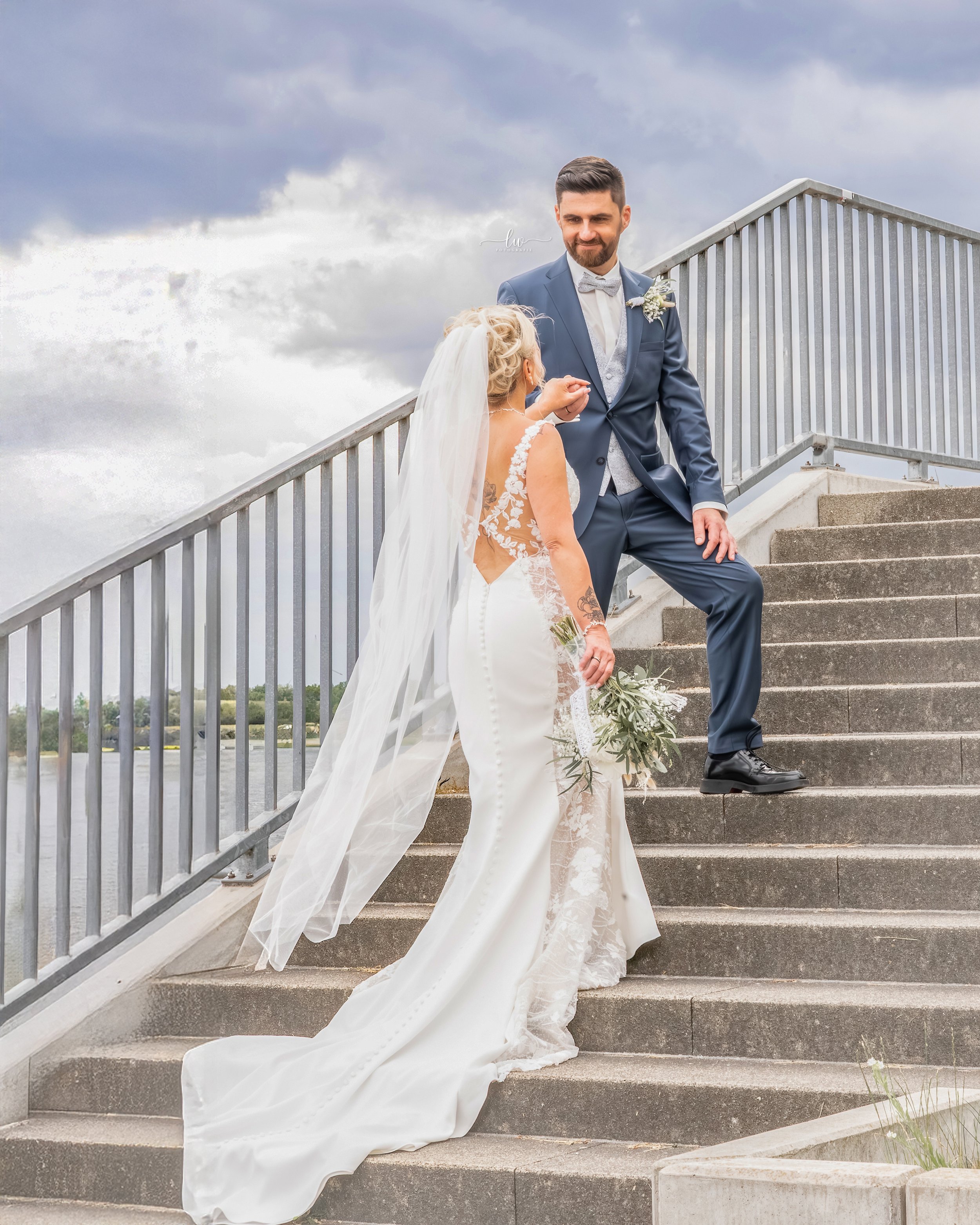 Ein Brautpaar auf einer Treppe im Freien, das Brautkleid und Anzug trägt, bei einer Hochzeitszeremonie, das Wetter ist bewölkt.