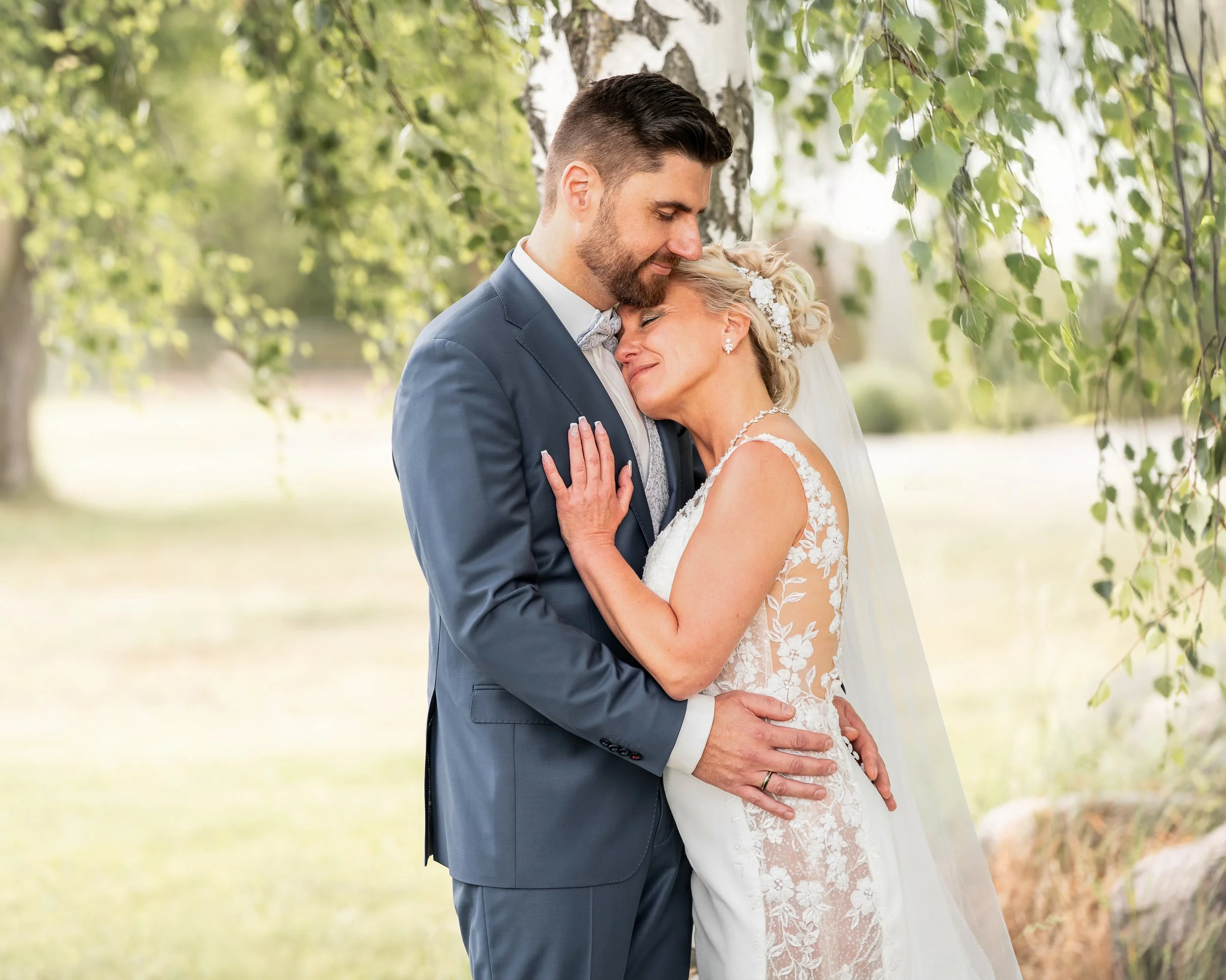 Ein Brautpaar in Abhängigkeit von nahegelegenen Bäumen bei Sonnenlicht, umarmend und einander liebend bei ihrer Hochzeit im Freien.