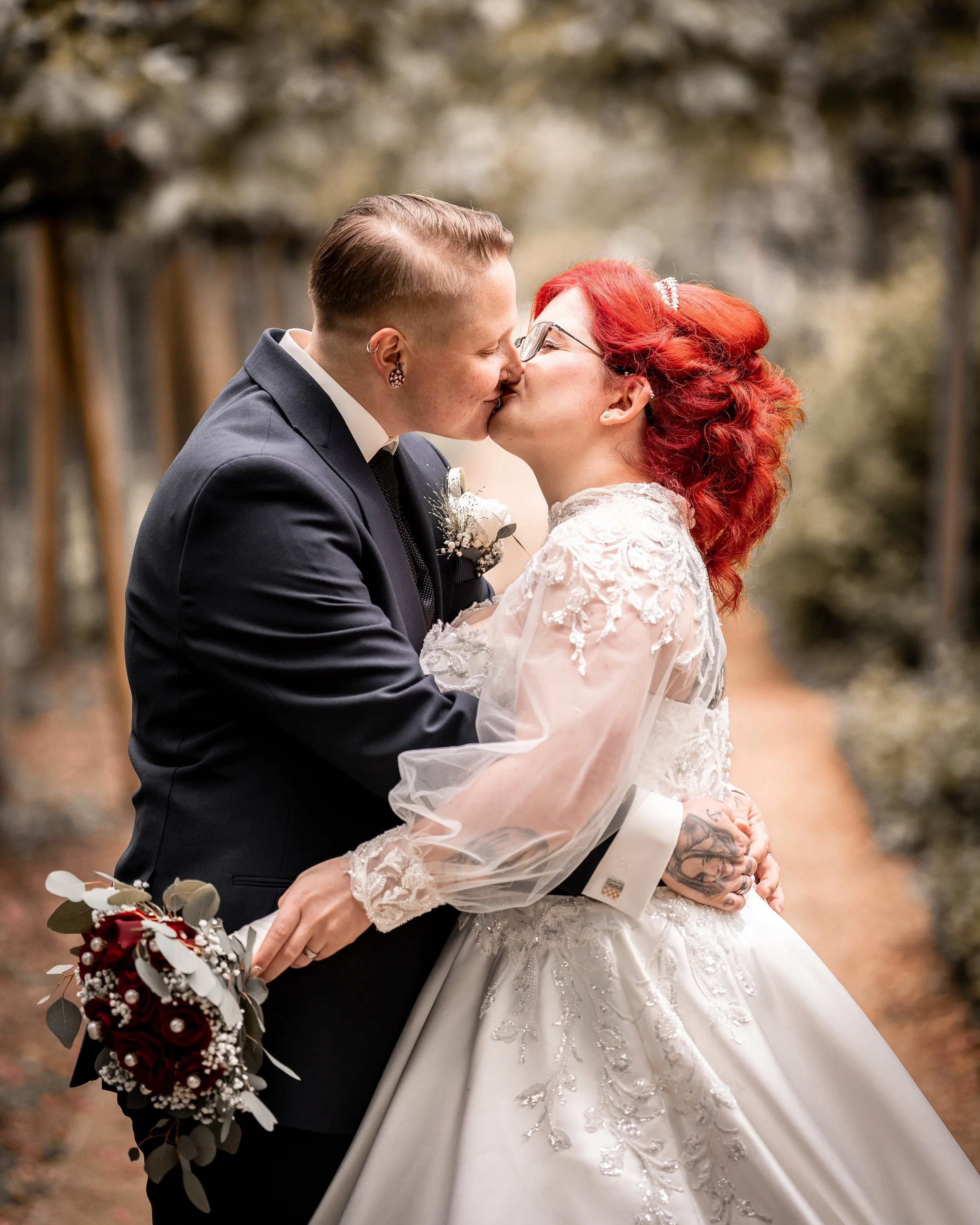 Ein Brautpaar küsst sich bei einer Hochzeit im Freien, die Frau hat rote Haare, trägt eine weiße Brautkleid mit verzierter Spitze, der Mann trägt einen schwarzen Anzug und eine Fliege, im Hintergrund sind Bäume.