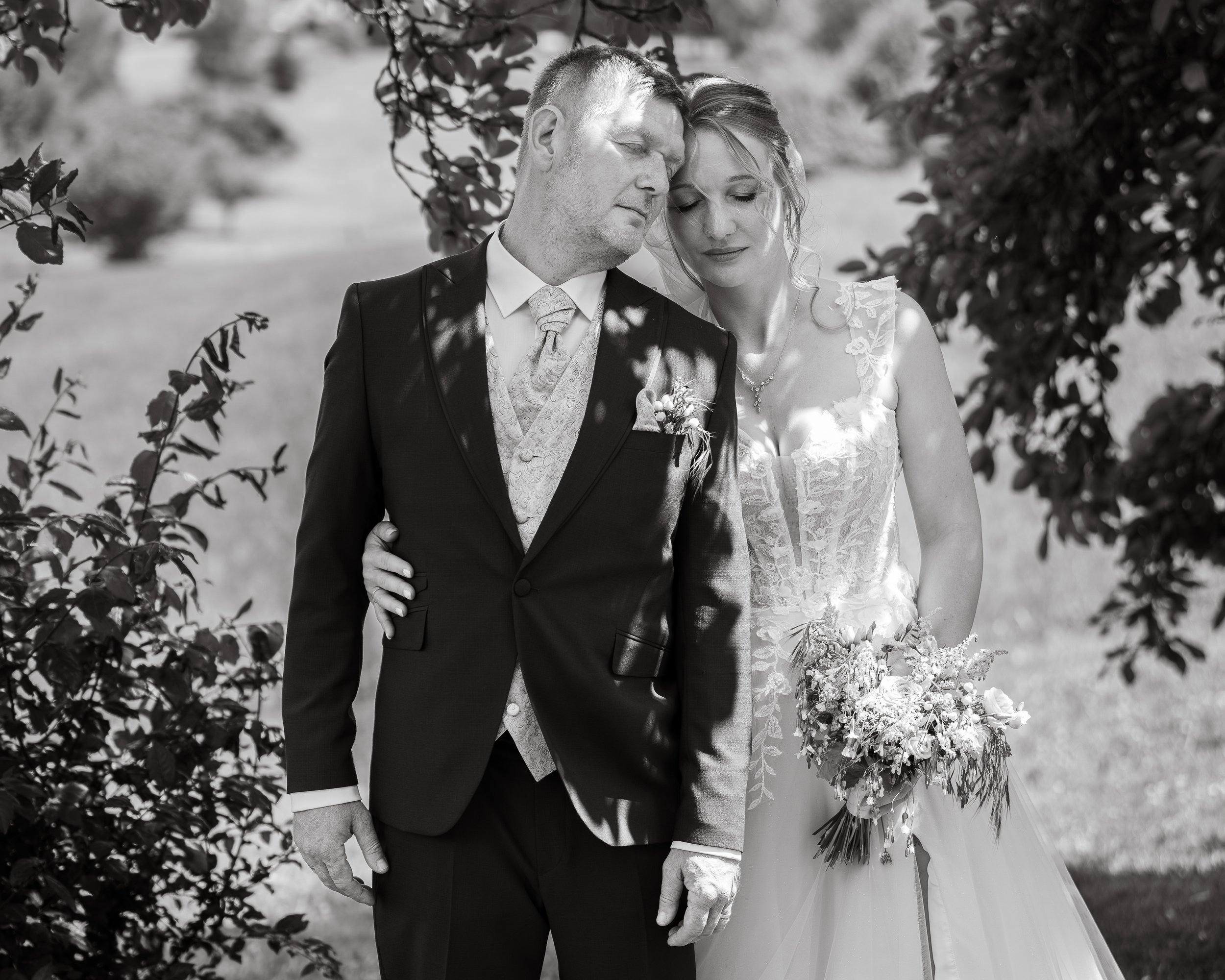Ein Mann und eine Frau bei einer Hochzeit, beide tragen formelle Kleidung, die Frau hält einen Blumenstrauß, sie stehen eng beieinander mit geschlossenen Augen, im Hintergrund sind Bäume und Zweige.