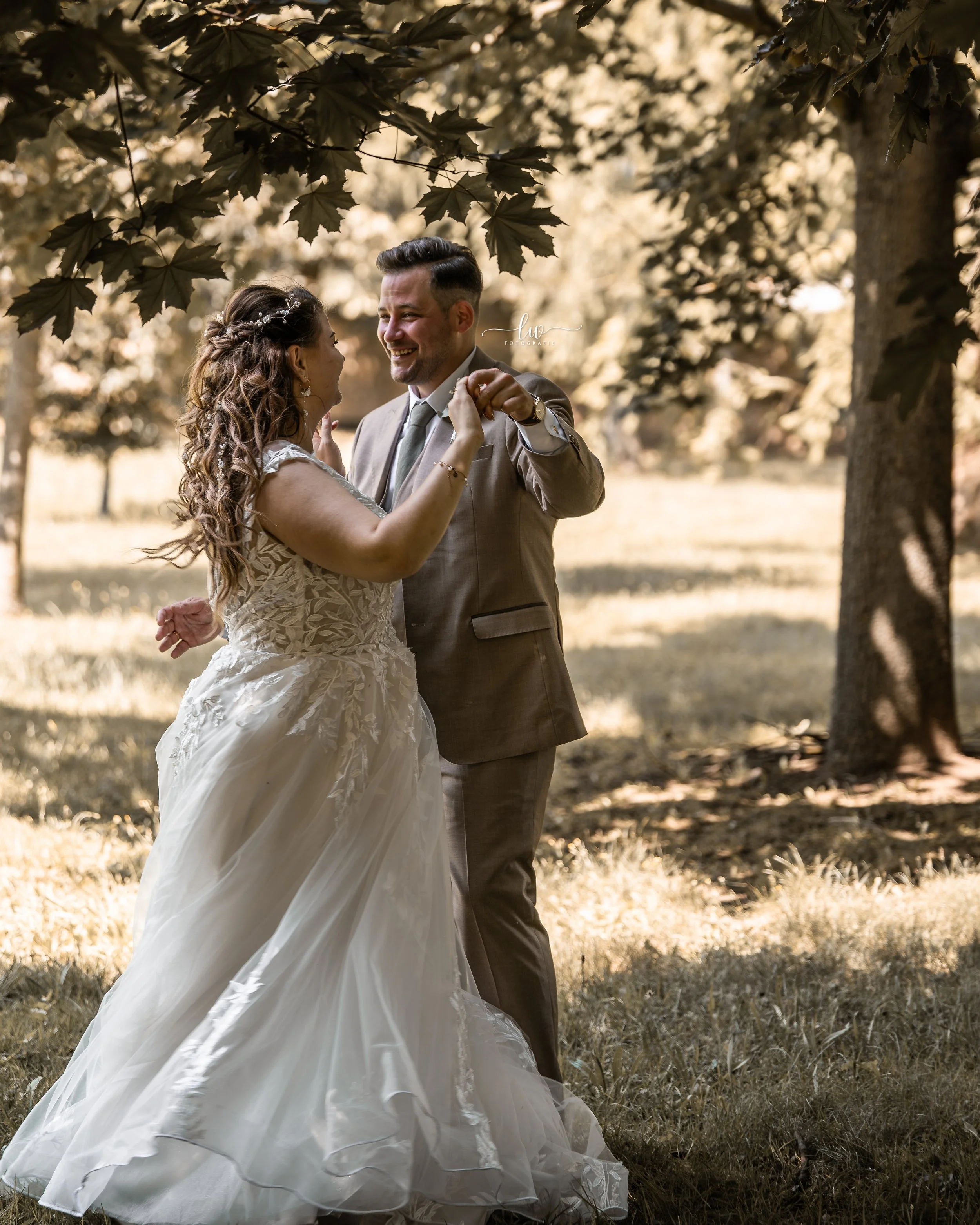 Ein Brautpaar tanzt unter Bäumen im Sonnenlicht, die Frau trägt ein elegantes Brautkleid mit Spitzenärmeln, der Mann einen Anzug mit Krawatte.