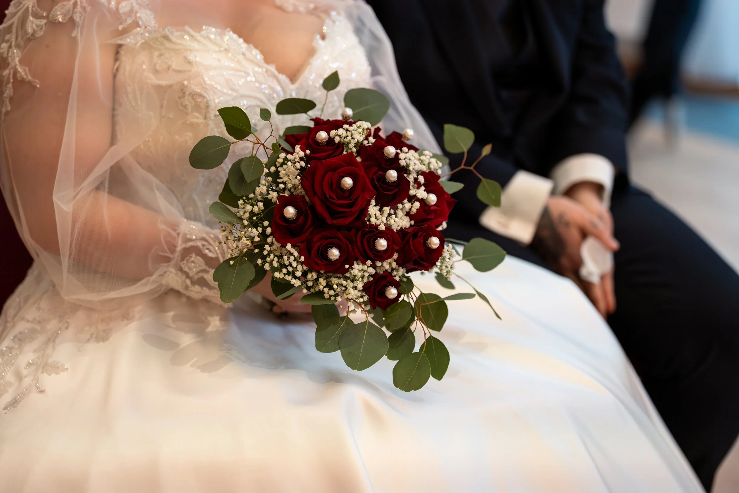 Hochzeitsstrauß mit roten Rosen, weißen Blümchen und grünen Blättern, gehalten von einer Braut in einem weißen Brautkleid. Im Hintergrund sitzen ein Brautpaar, der Bräutigam in Schwarz und die Braut in Weiß.