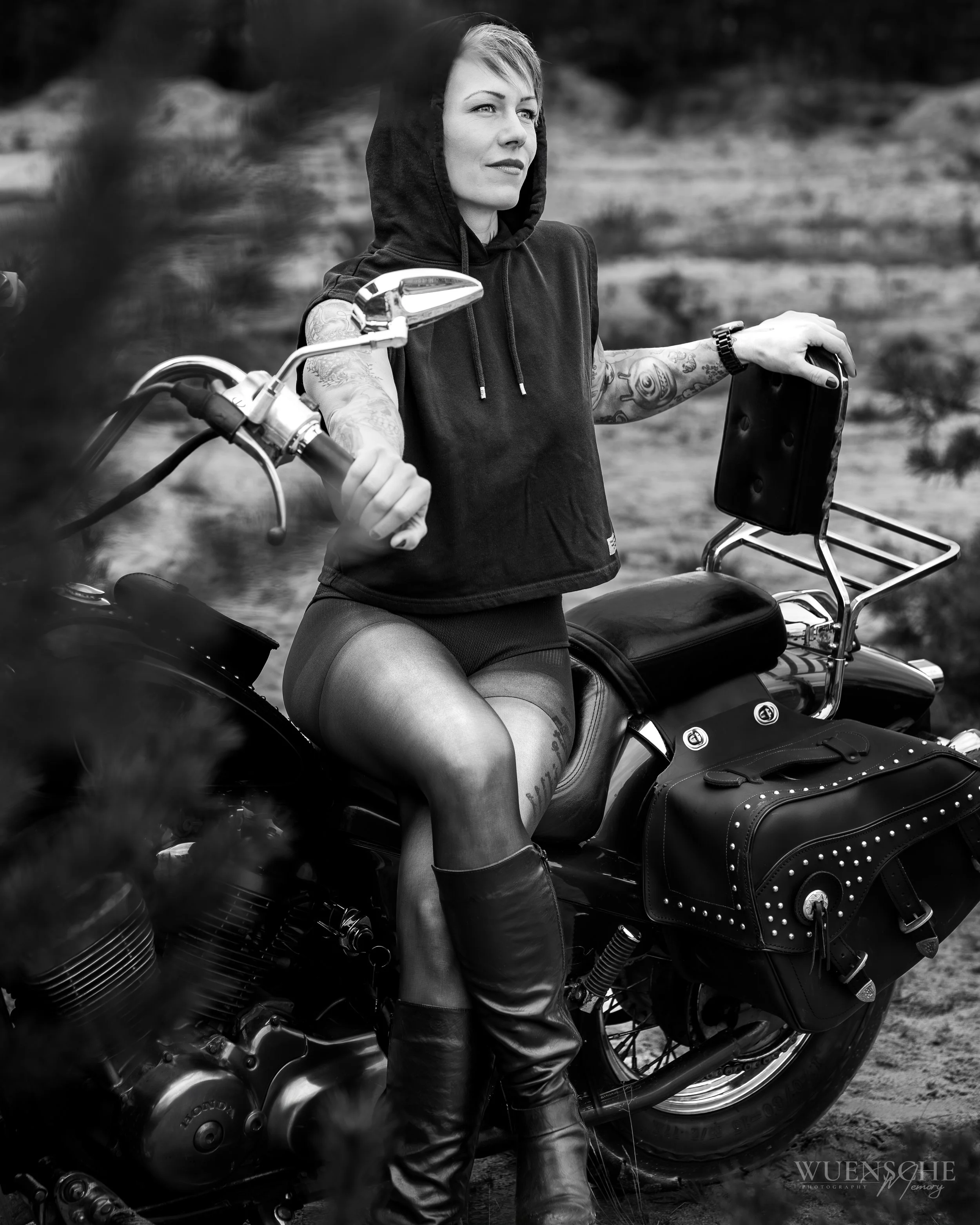 Schwarz-weiß-Fotografie einer jungen Frau mit Tattoos auf den Armen, die auf einem Motorrad sitzt, draußen, mit Kapuzenpullover.
