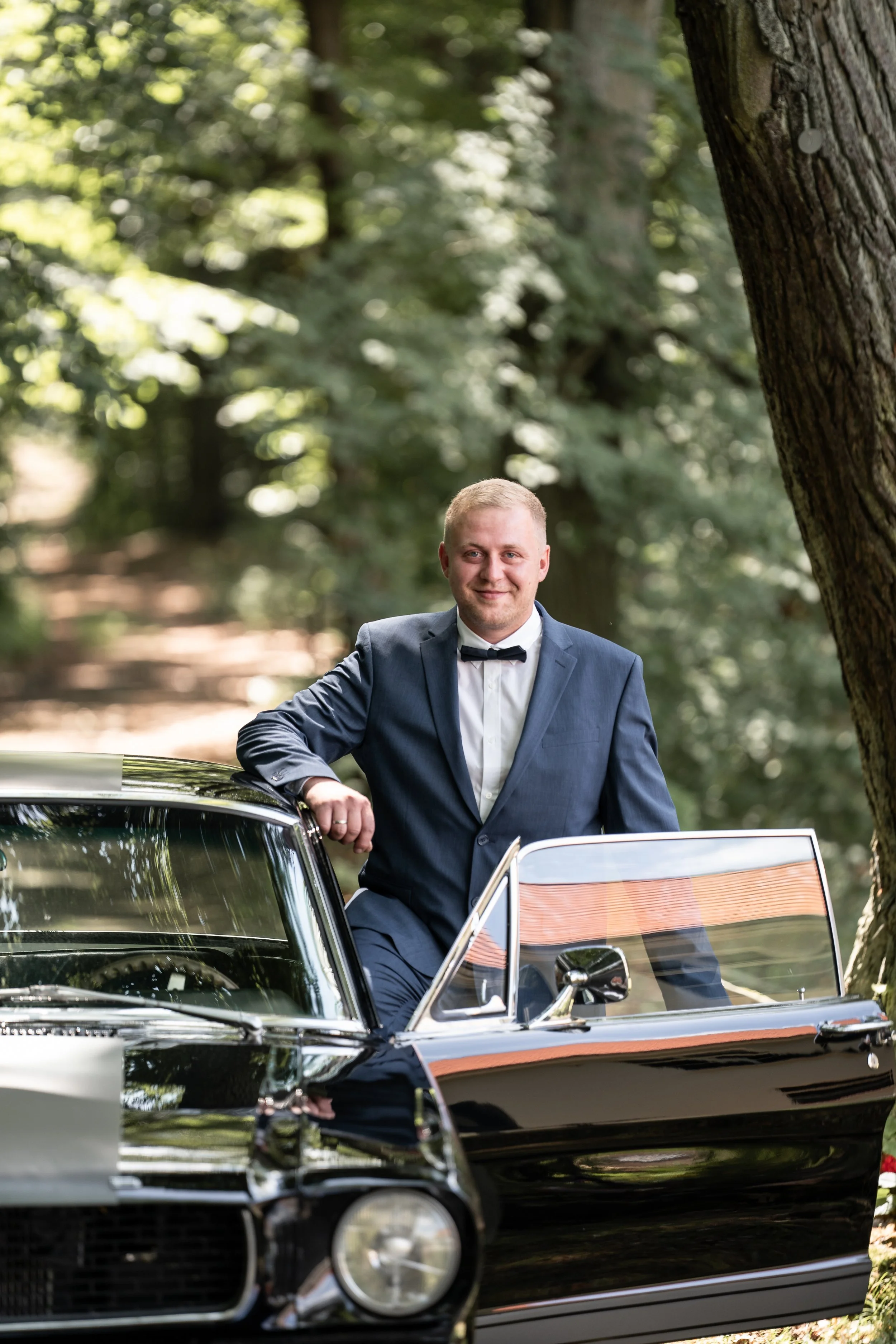 Junger Mann in Anzug mit Fliege steht neben einem klassischen schwarzen Auto im Wald.