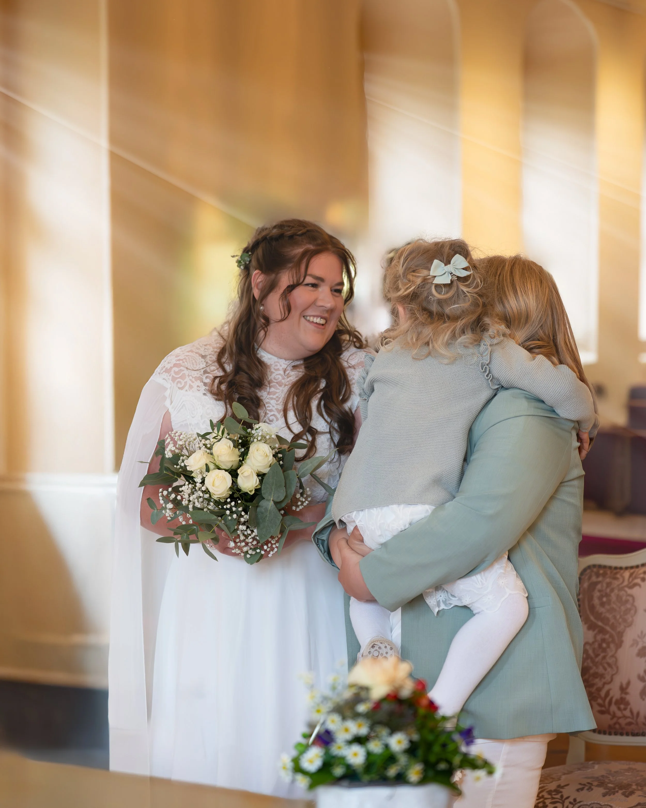 Eine Frau in einem weißen Hochzeitskleid hält ein Brautstrauß und lächelt eine kleine blonde Mädchen an, während sie eine Umarmung austauschen. Die Szene findet wahrscheinlich bei einer Hochzeit statt, in einem hell erleuchteten Raum mit warmen Farbtönen.