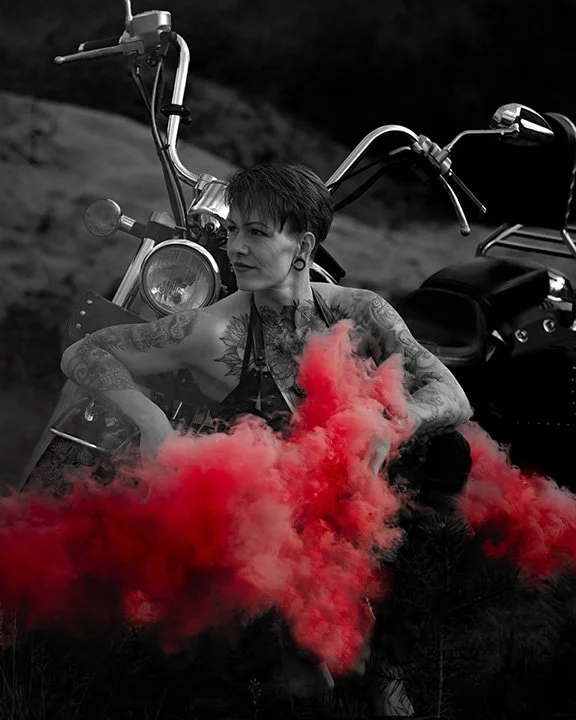 Schwarz-weiß-Foto eines tattoosierten Mannes mit kurzen schwarzen Haaren, der vor einem Motorrad sitzt und rote Rauchwolken um ihn herum aufsteigt.