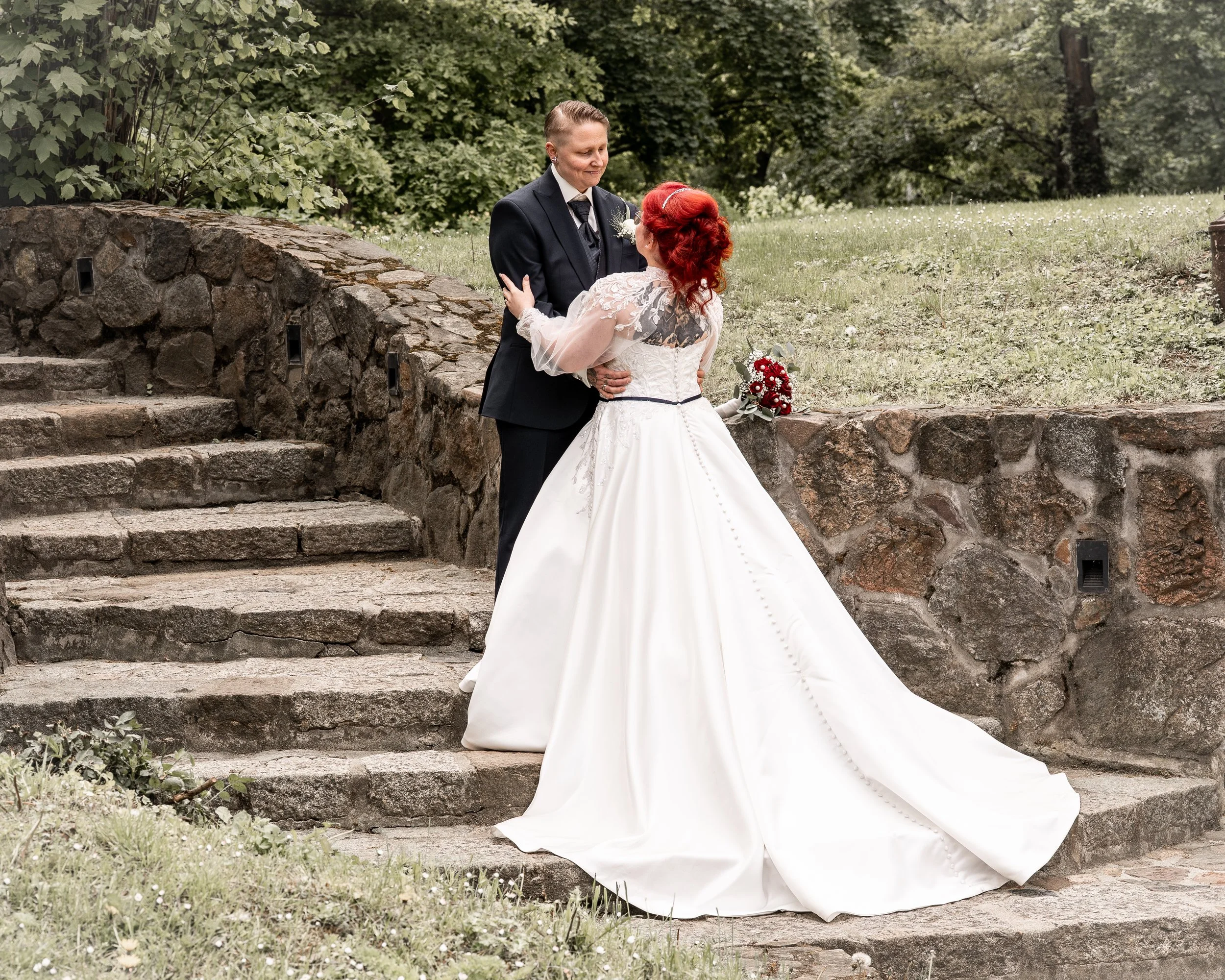 Ein Brautpaar steht vor einer Steinmauer im Freien, die Braut trägt ein weißes Hochzeitskleid mit Spitze und rot gefärbte Haare, der Bräutigam trägt einen schwarzen Anzug mit Krawatte, im Hintergrund sind grüne Bäume.