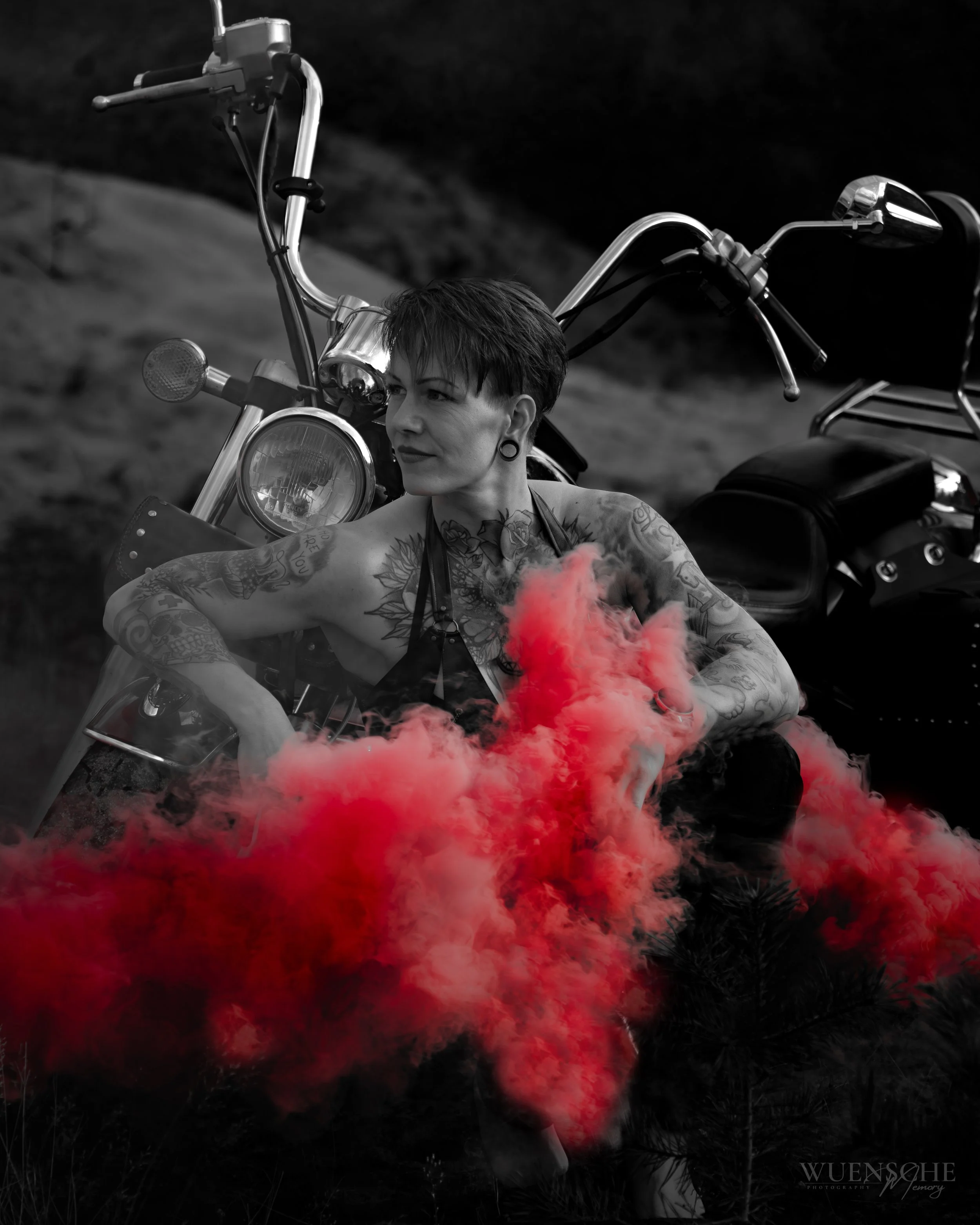 Schwarze und weiße Fotografie eines Motorrads mit einem Gesicht einer Frau mit Tattoos im Vordergrund, umgeben von roten Rauchwolken, im Dunkeln.