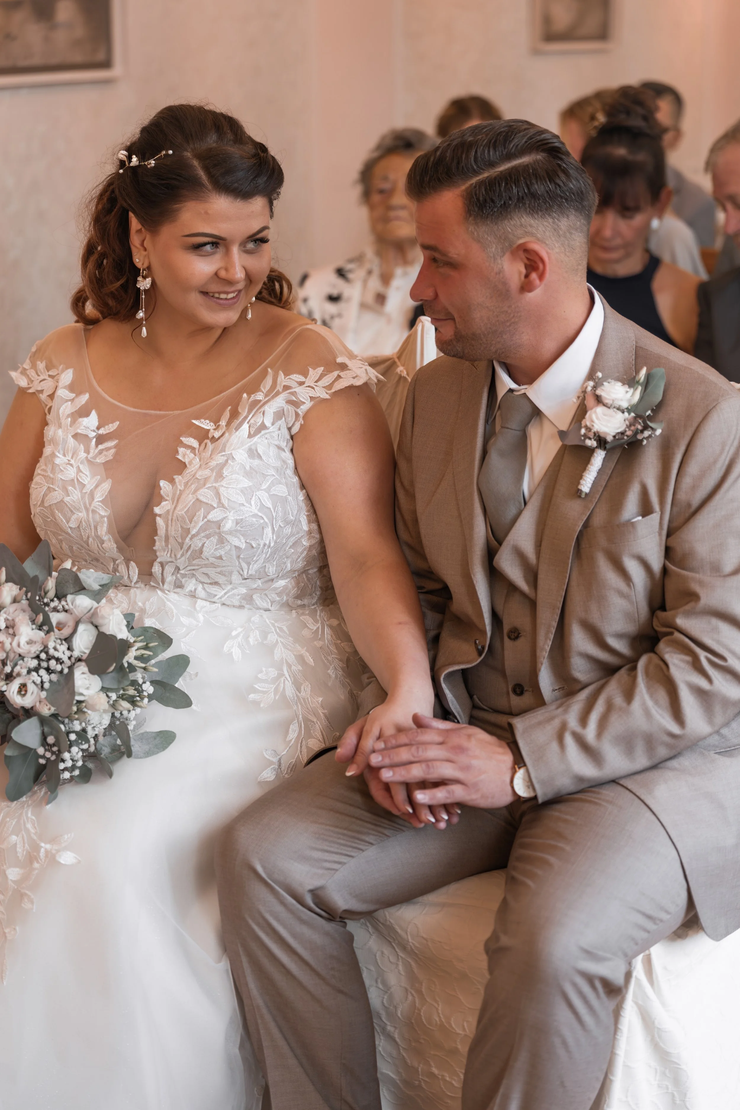 Ein Brautpaar sitzt während ihrer Hochzeitszeremonie, die Braut trägt ein weißes Kleid mit Spitzen und Perlenohrringen, der Bräutigam trägt einen beigen Anzug mit einer Blume im Revers. Sie halten Händchen, im Hintergrund stehen Gäste in verschiedenen Outfits.