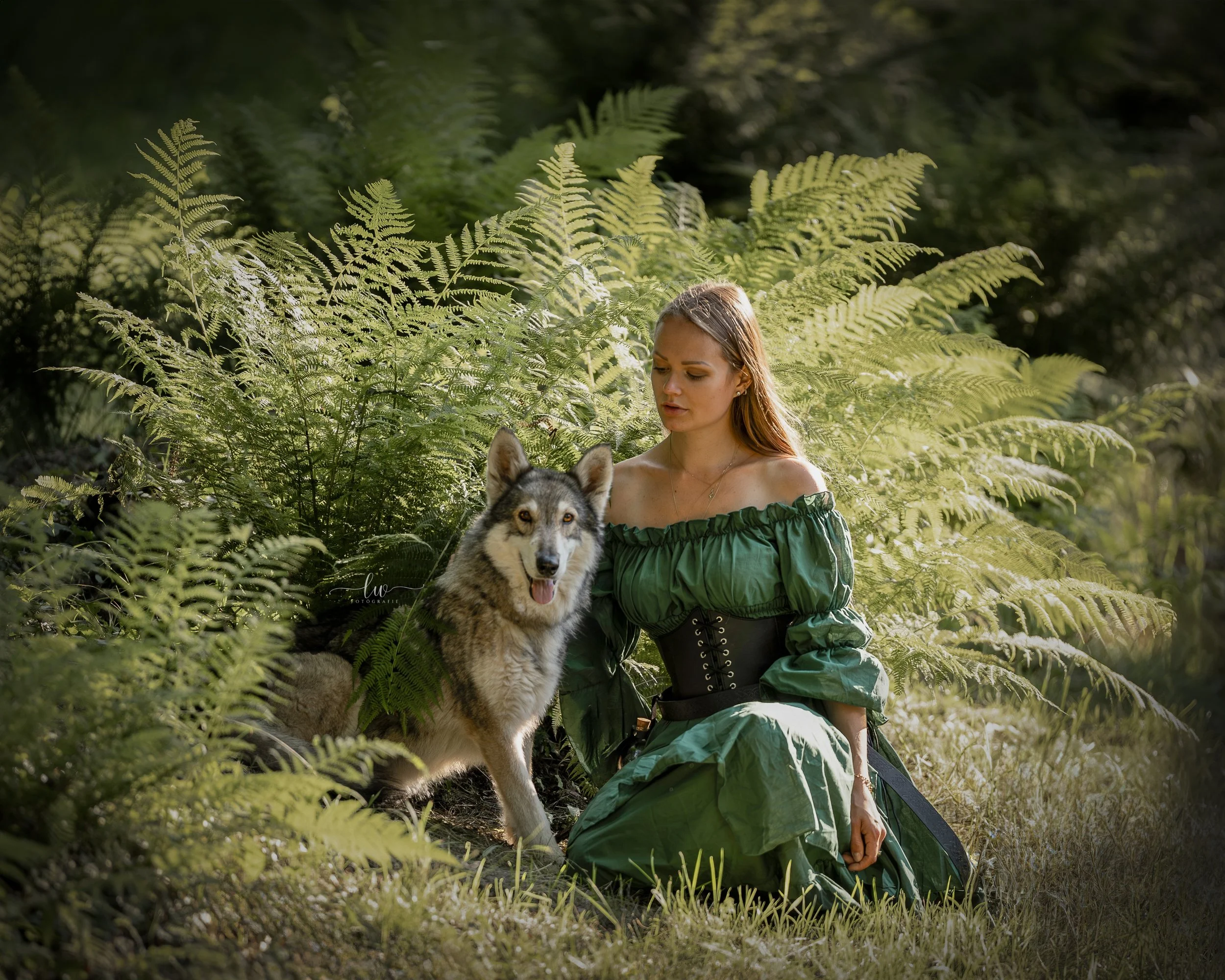 Junge Frau mit langem Haar in grüner Kleidung sitzt im Wald neben einem Hund, umgeben von hohen Farnen und dichtem Grün, mit Sonnenlicht, das durch die Blätter fällt.