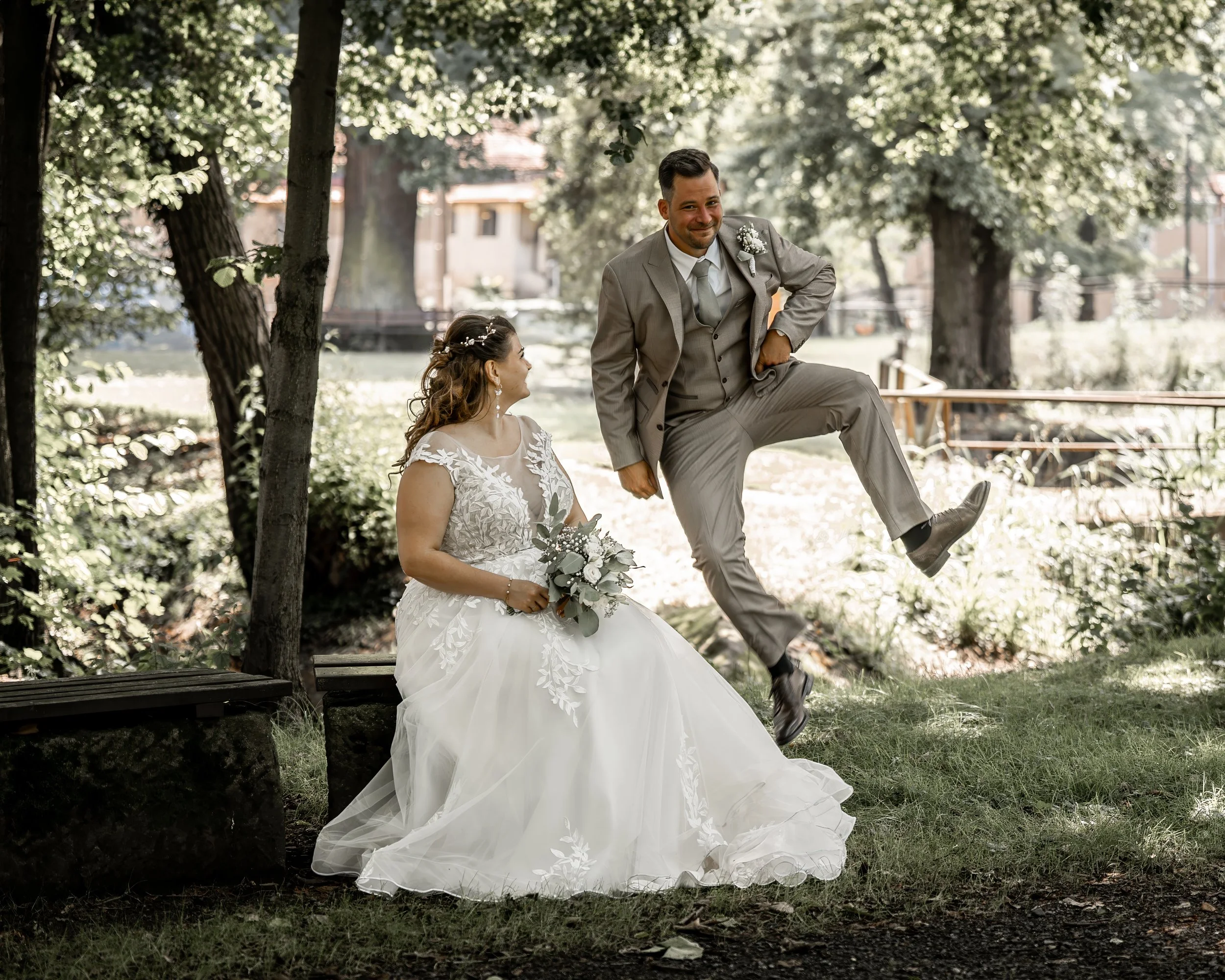Braut und Bräutigam im Park, der Bräutigam sitzt auf einem Baumstamm mit Anzug und Lächeln, die Braut sitzt auf dem Boden in einem weißen Brautkleid mit Blumen im Haar und hält einen Blumenstrauß, umgeben von Bäumen und Sonnenlicht.