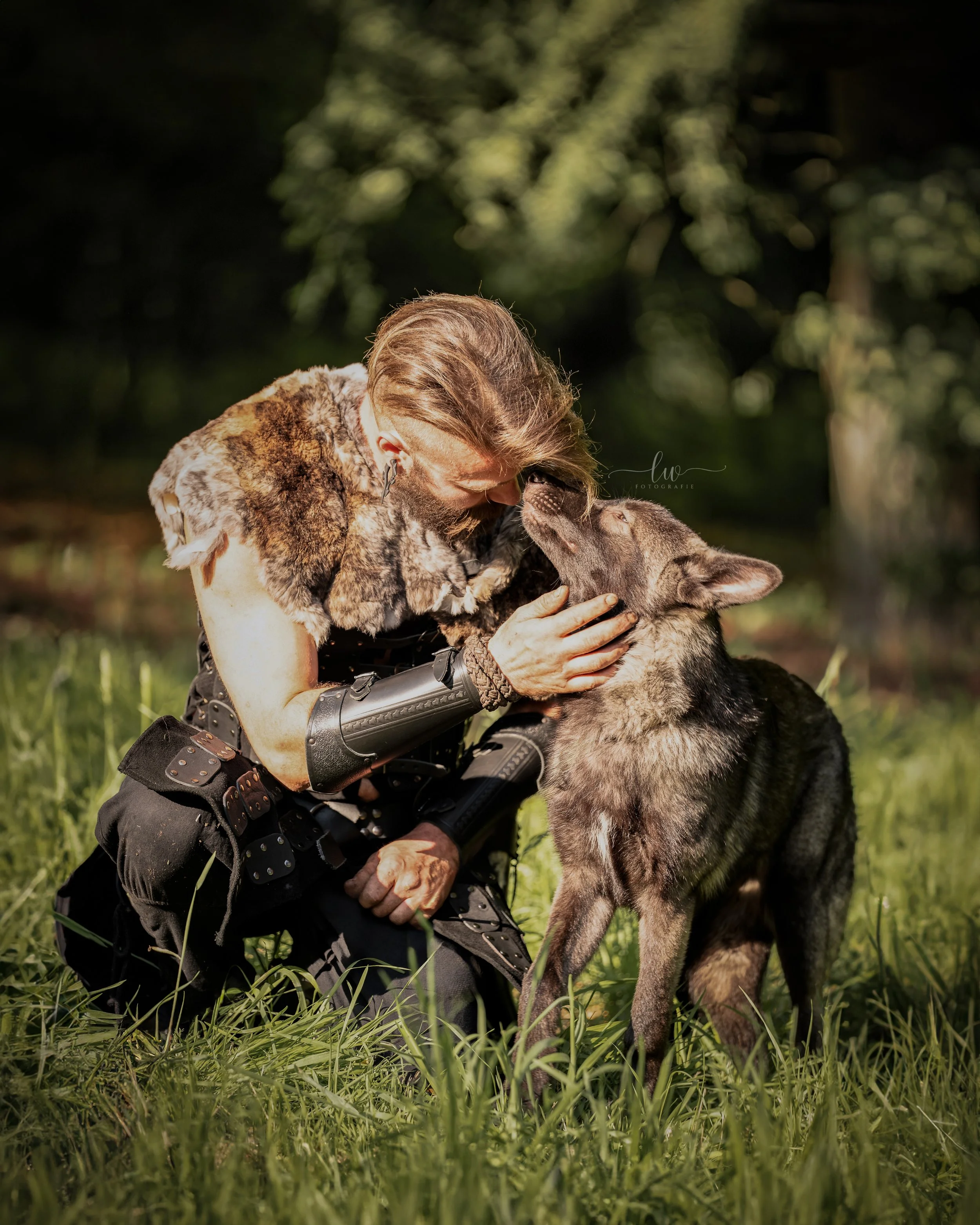 Ein Mann mit einem künstlichen Arm hält ein wolfähnliches Tier im Grase auf einer Wiese, umgeben von Bäumen.