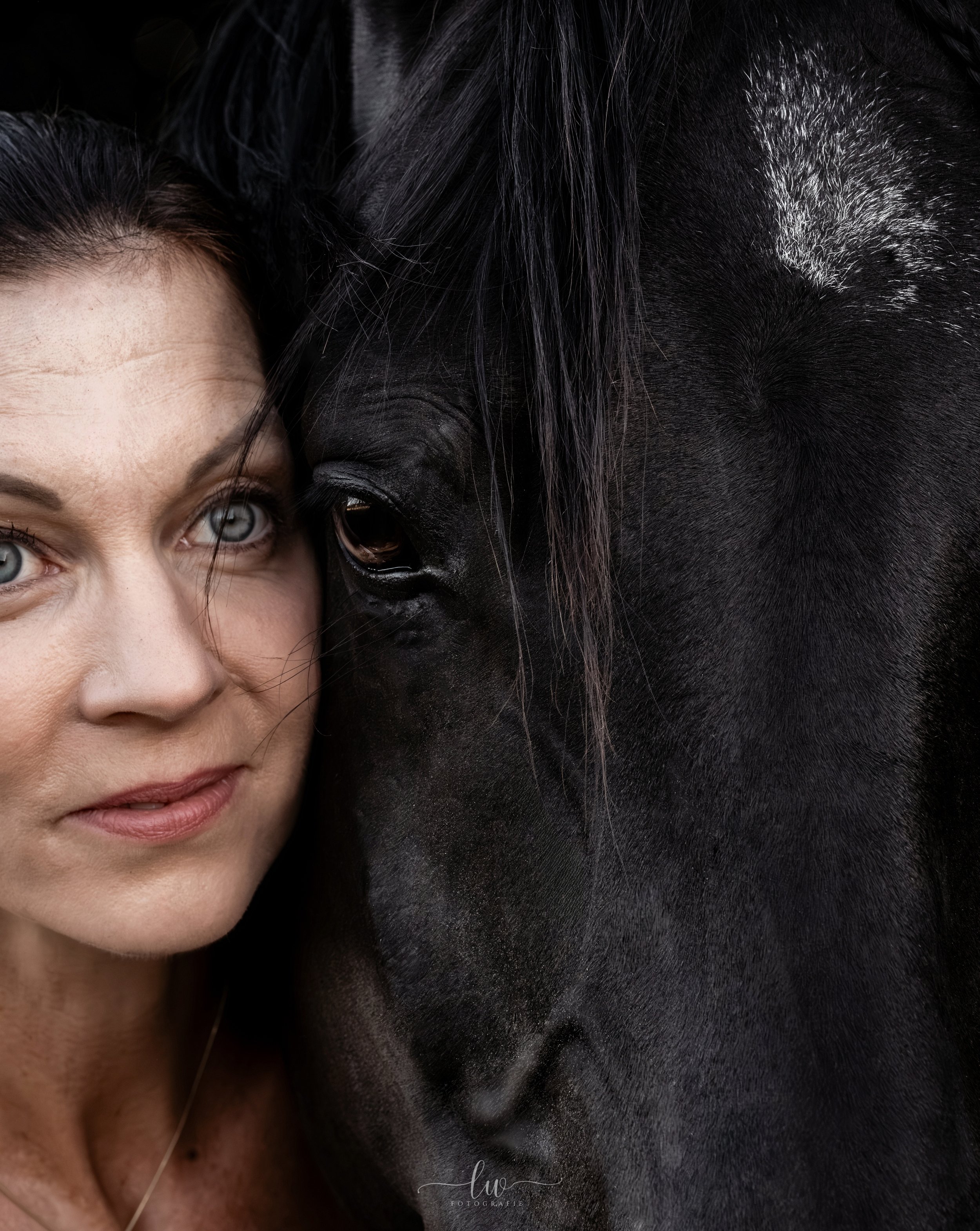 Nahaufnahme einer Frau mit blauen Augen und dunklen Haaren, die neben einem schwarzen Pferd steht.