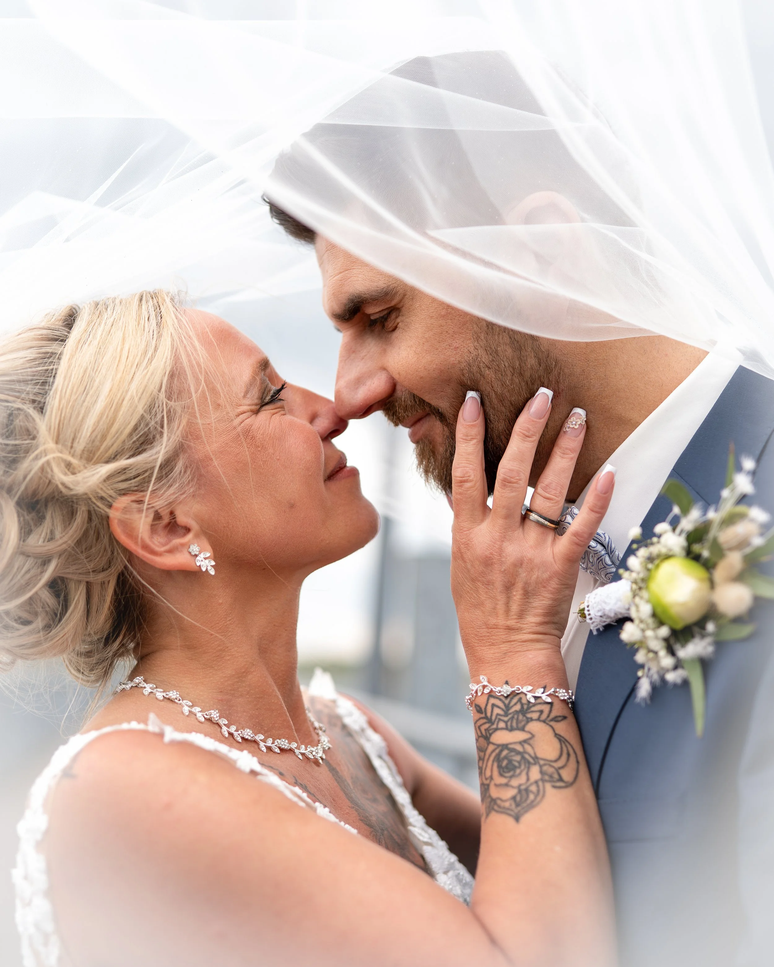Ein Brautpaar bei einer Hochzeitszeremonie, sie stehen sich sehr nah, mit dem Blick in die Augen, unter einem weißen Schleier, die Frau lächelt, trägt geschmückten Schmuck, der Mann hat Tattoos und trägt einen Blumenstrauß