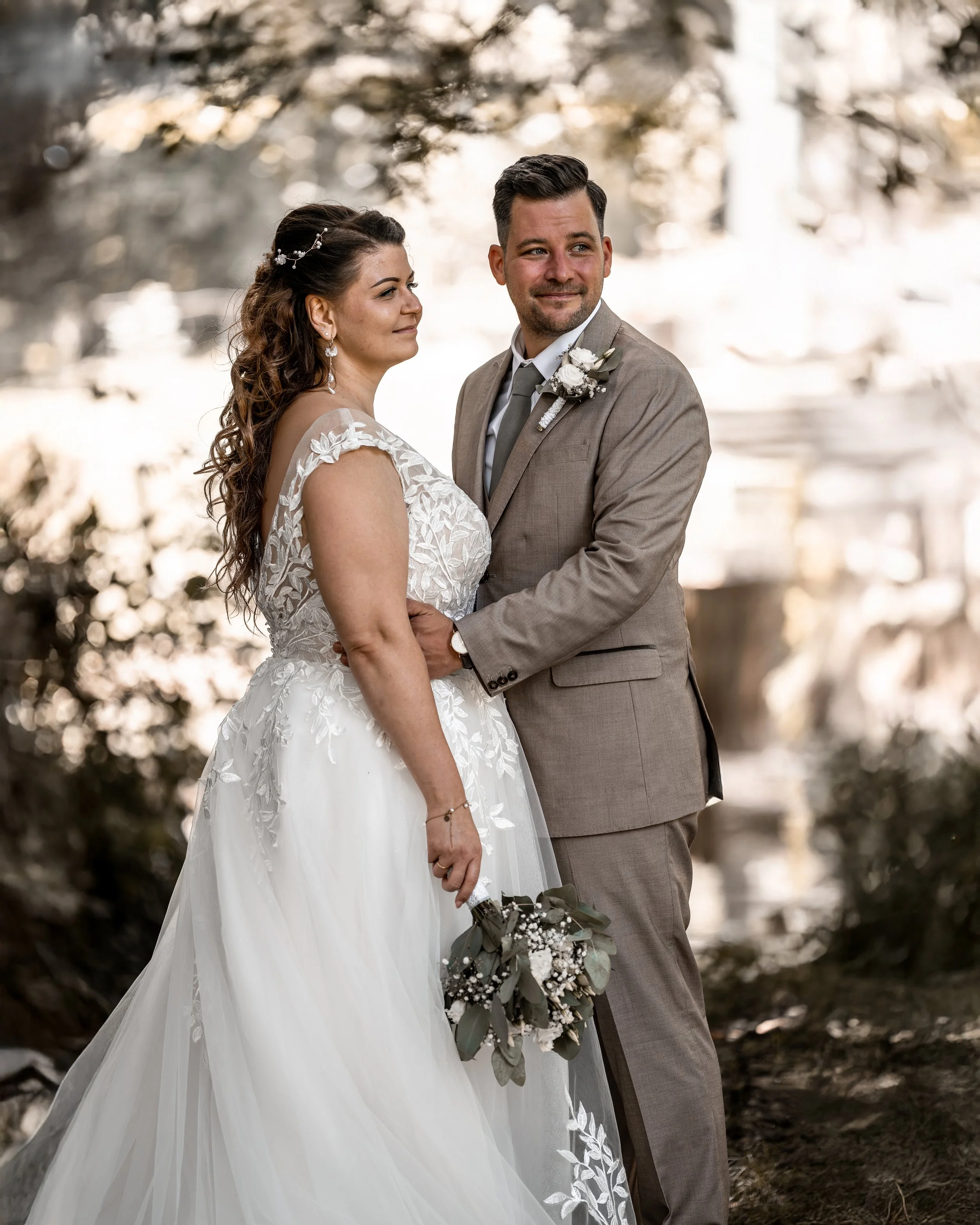 Ein Brautpaar bei einer Hochzeit im Freien, die Braut trägt ein weißes Kleid mit Spitzen und Blumenmuster, der Bräutigam trägt einen hellbraunen Anzug mit Blume im Revers, sie halten Händchen, im Hintergrund Bäume mit Blättern und Sonnenlicht