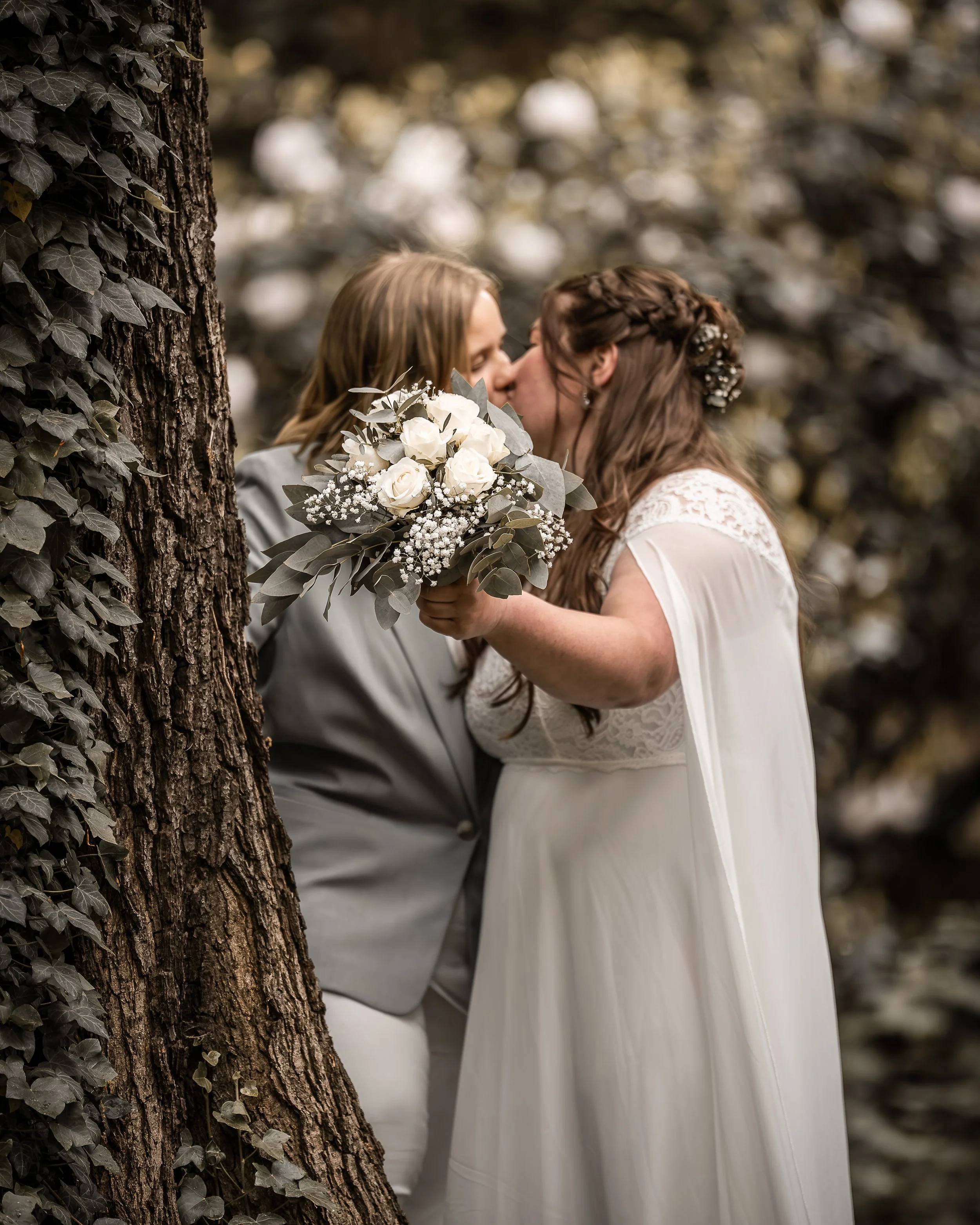 Zwei Frauen küssen sich bei einer Hochzeit im Freien, eine hält einen Brautstrauß aus weißen Rosen und Blättern, im Hintergrund Blätter und Bäume.