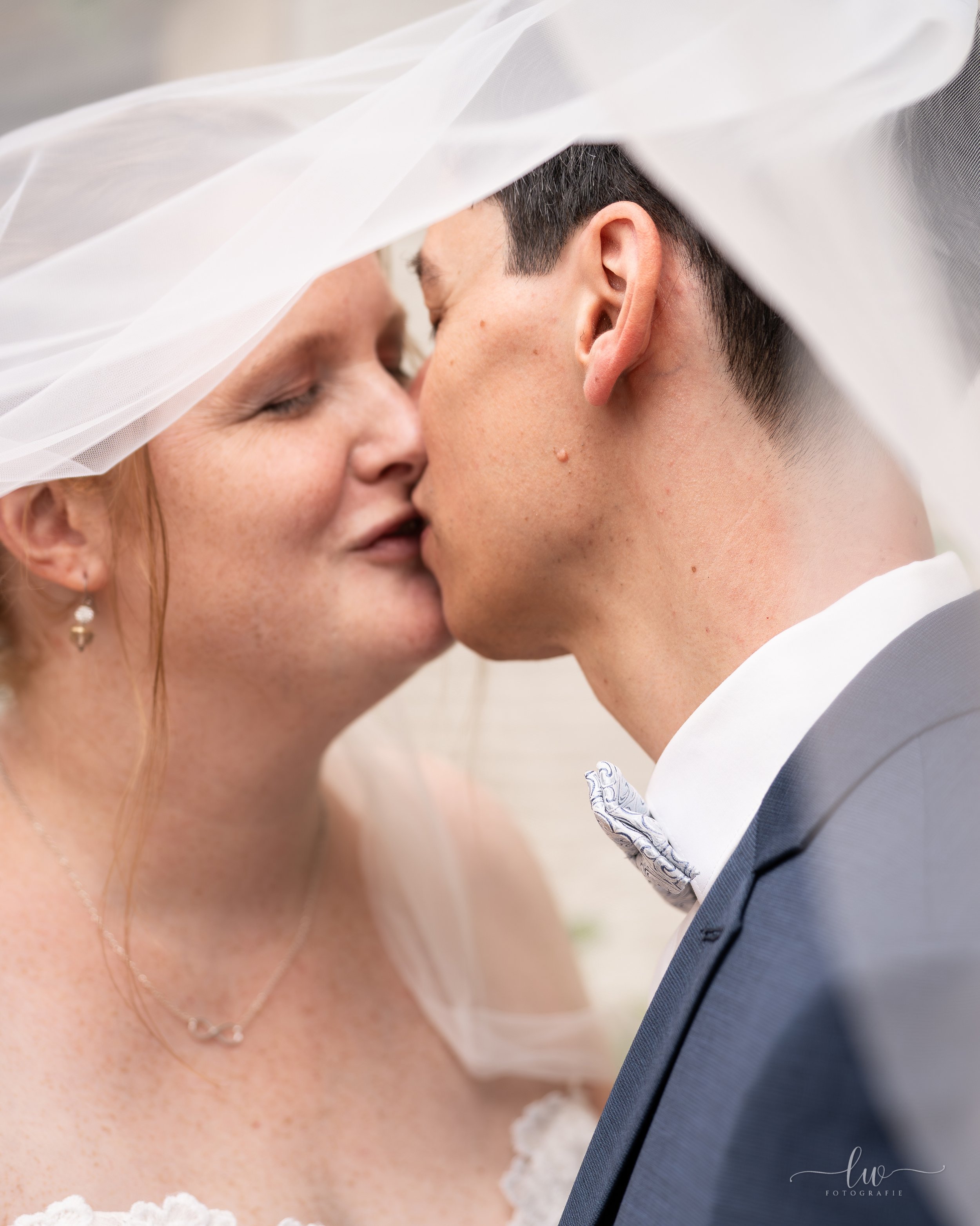 Ein Brautpaar berührt sich mit der Stirn, die Braut trägt eine Tüll-Haarschleife und einen weißen Hochzeitskleid, der Bräutigam trägt einen dunklen Anzug. Sie sind sehr emotional und romantisch bei ihrer Hochzeitsfeier.