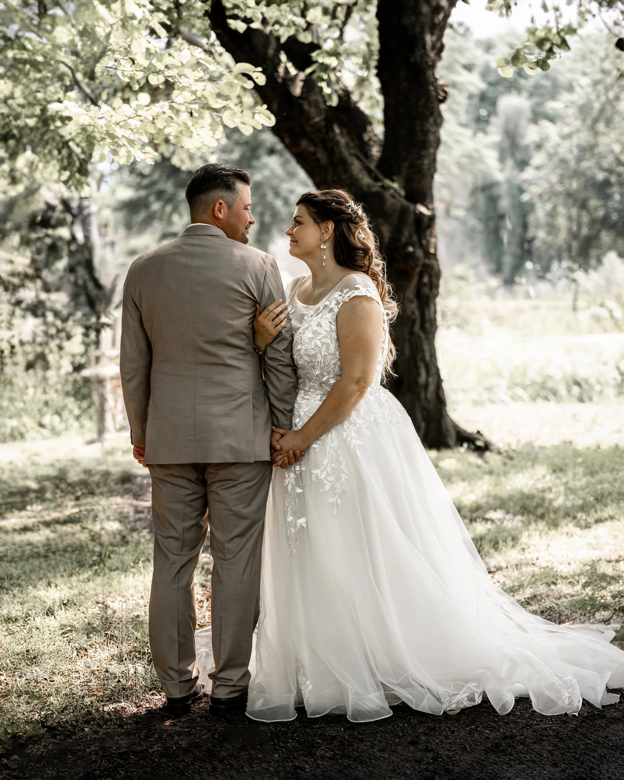 Ein Brautpaar steht sich in einem Wald mit leuchtend grünen Bäumen gegenüber. Sie lächeln sich an, die Frau trägt ein weißes Brautkleid mit floralen Details und eleganten Ohrringen, der Mann trägt einen grauen Anzug. Die Szenerie ist sonnig und die Atmosphäre ist romantisch.