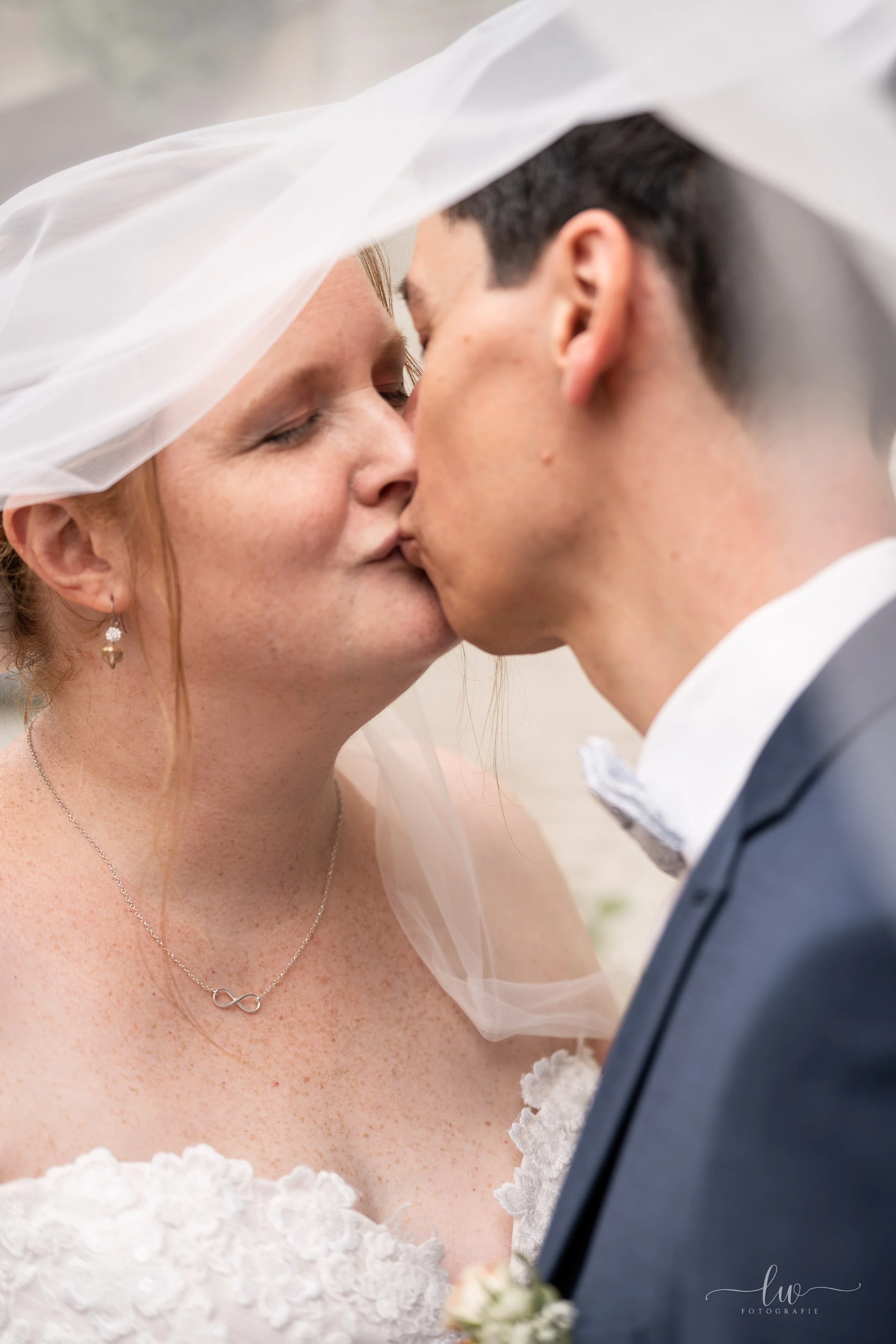 Braut und Bräutigam küssen sich an ihrer Hochzeit, die Braut trägt eine weiße Hochzeitstracht und eine Kette mit Unendlichkeitssymbol, der Bräutigam trägt einen dunklen Anzug und Fliege.