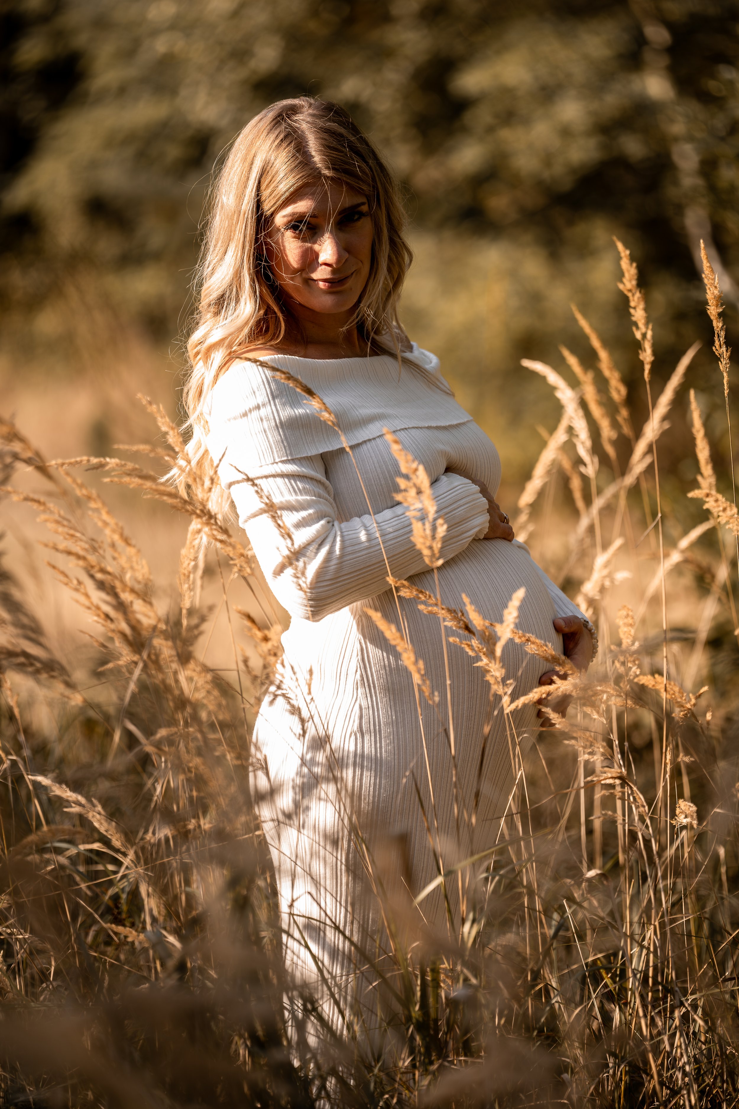 Schwangere Frau steht in einem Feld mit goldenen Gräsern bei Sonnenuntergang.